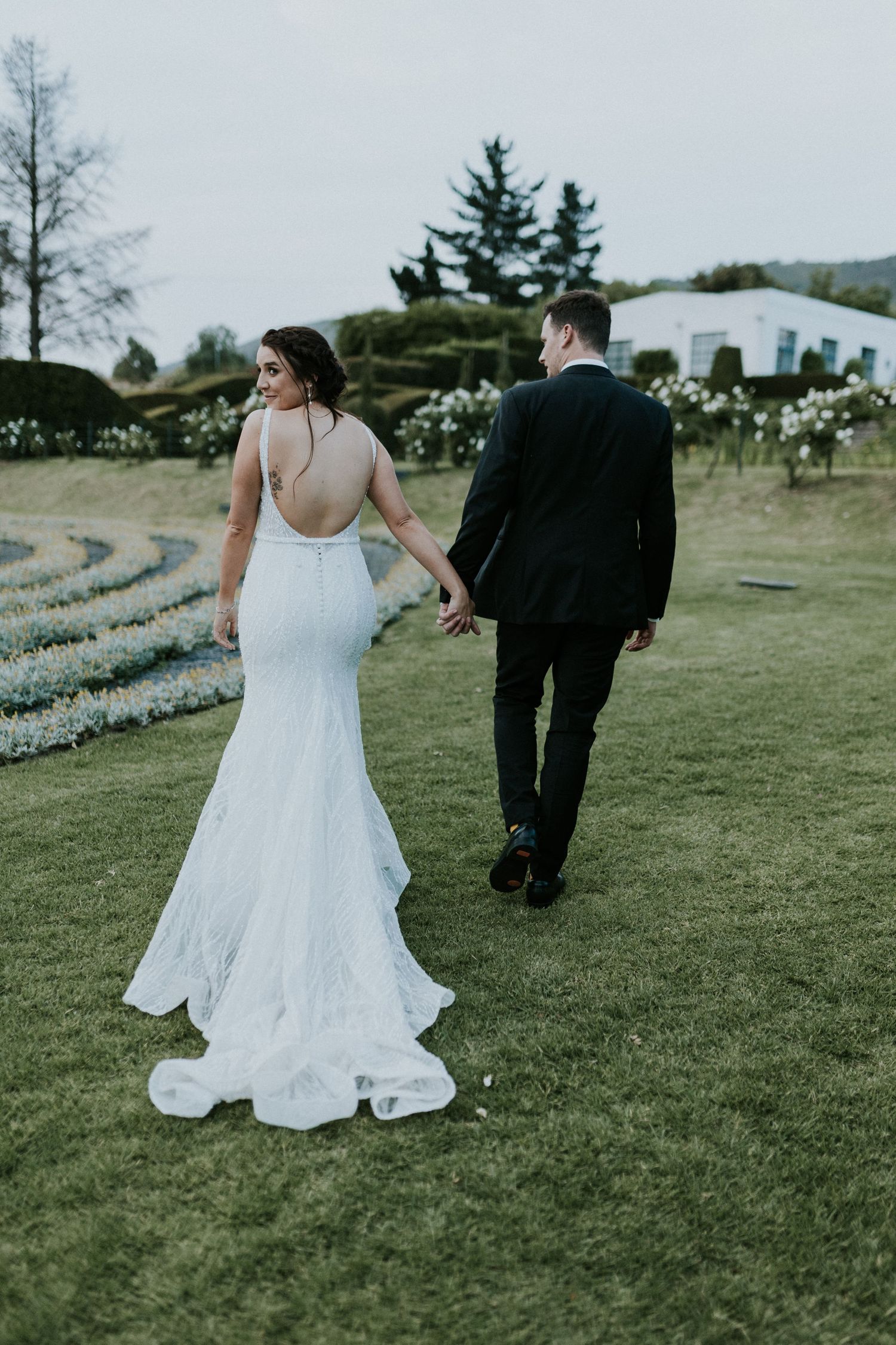 Bride in backless mermaid wedding gown holds groom's hand walking through manicured garden grounds.
