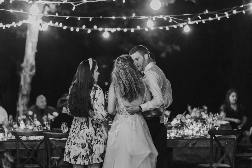Black and white photo of bride and groom sharing a dance with a guest under string lights.