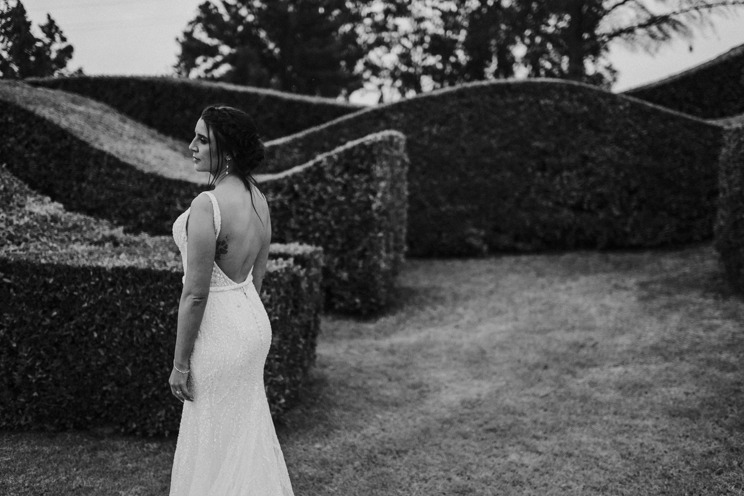 Black and white portrait of bride in backless wedding gown standing before sculpted topiary hedges.