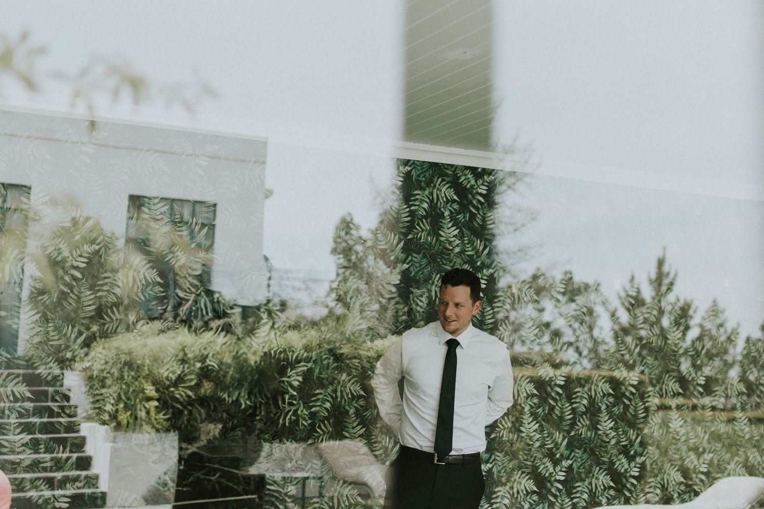 Groom in white shirt and black tie stands outdoors among lush greenery.