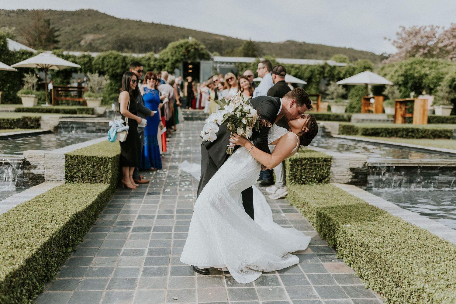 Groom dips his bride for a romantic kiss along a manicured garden pathway following their outdoor wedding ceremony.