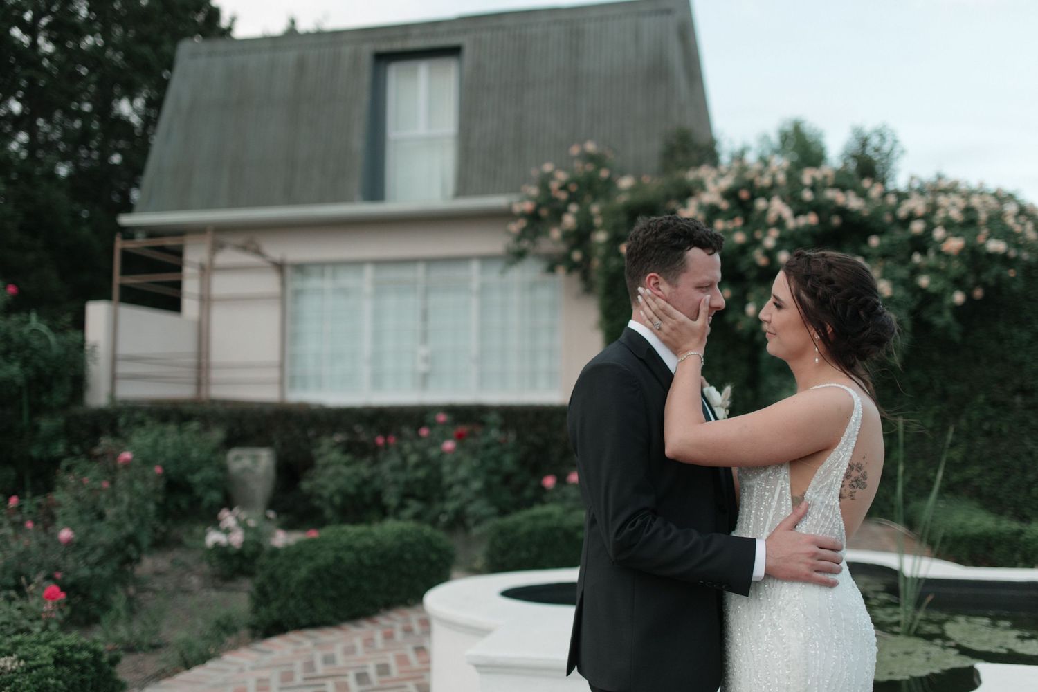 Bride and groom sharing a romantic moment in garden with rose bushes and charming house backdrop.