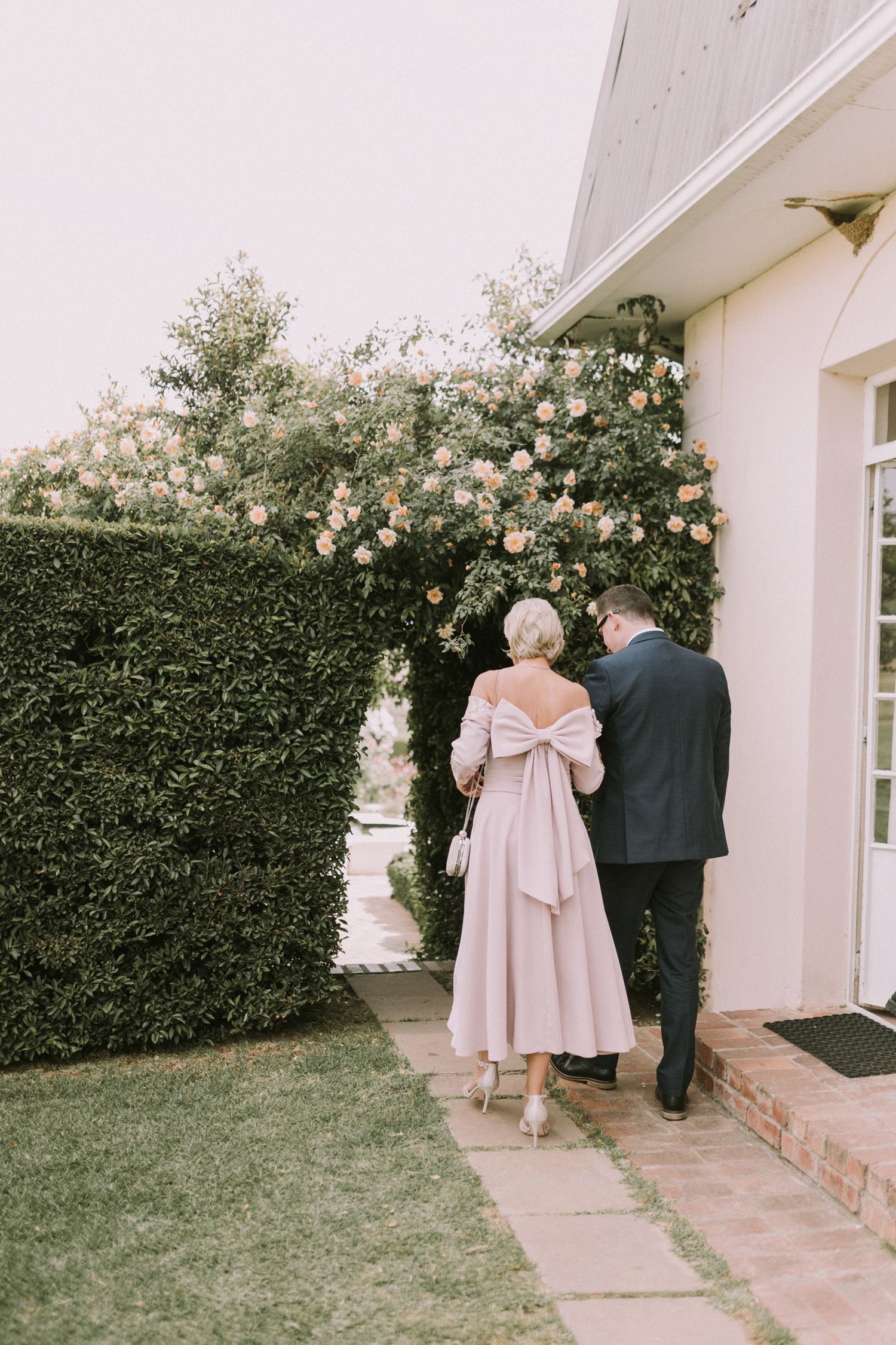 Woman in blush pink off-shoulder bow dress walking with suited man past blooming rose hedge.