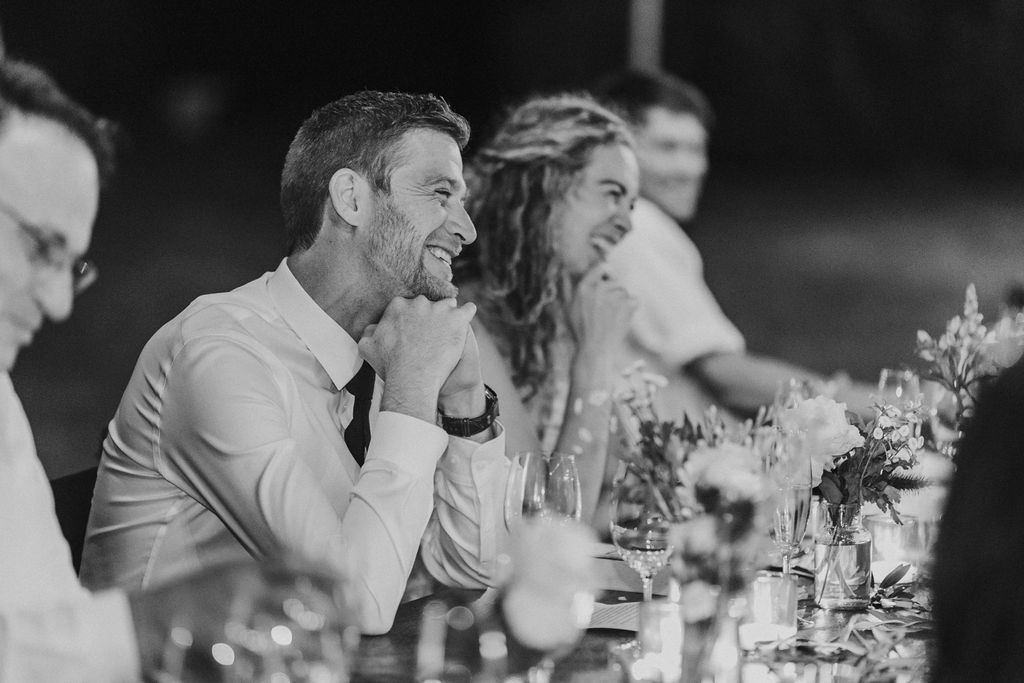 Handsome groom laughs heartily while seated at elegant wedding reception dinner table.