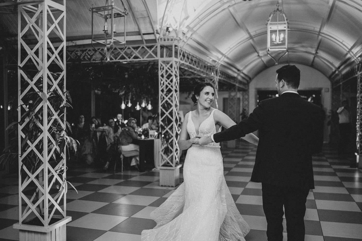 Black and white photo of bride and groom sharing first dance on checkered floor in ornate venue.