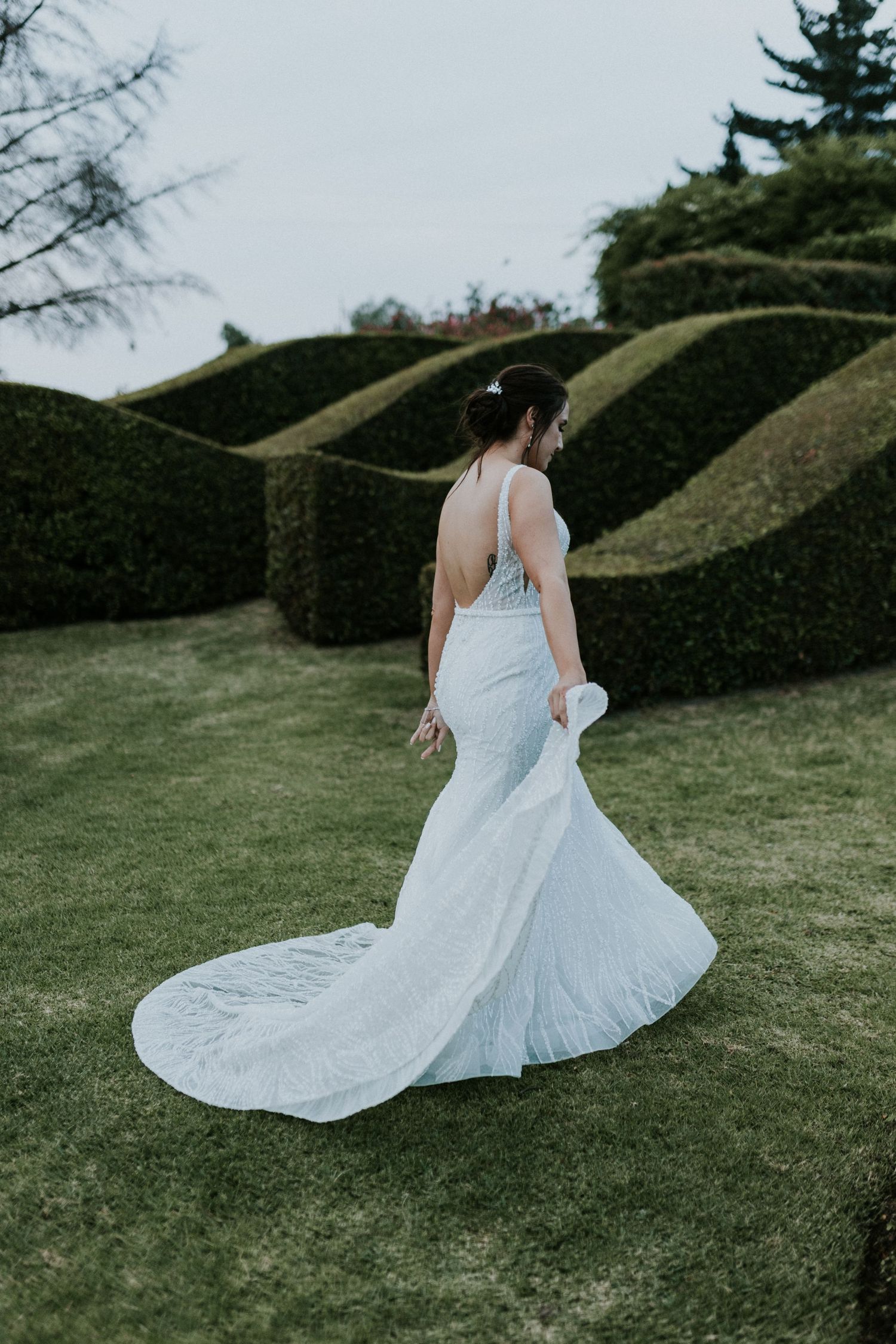 Bride in fitted mermaid wedding gown poses before dramatic wave-cut topiary hedges on manicured lawn.