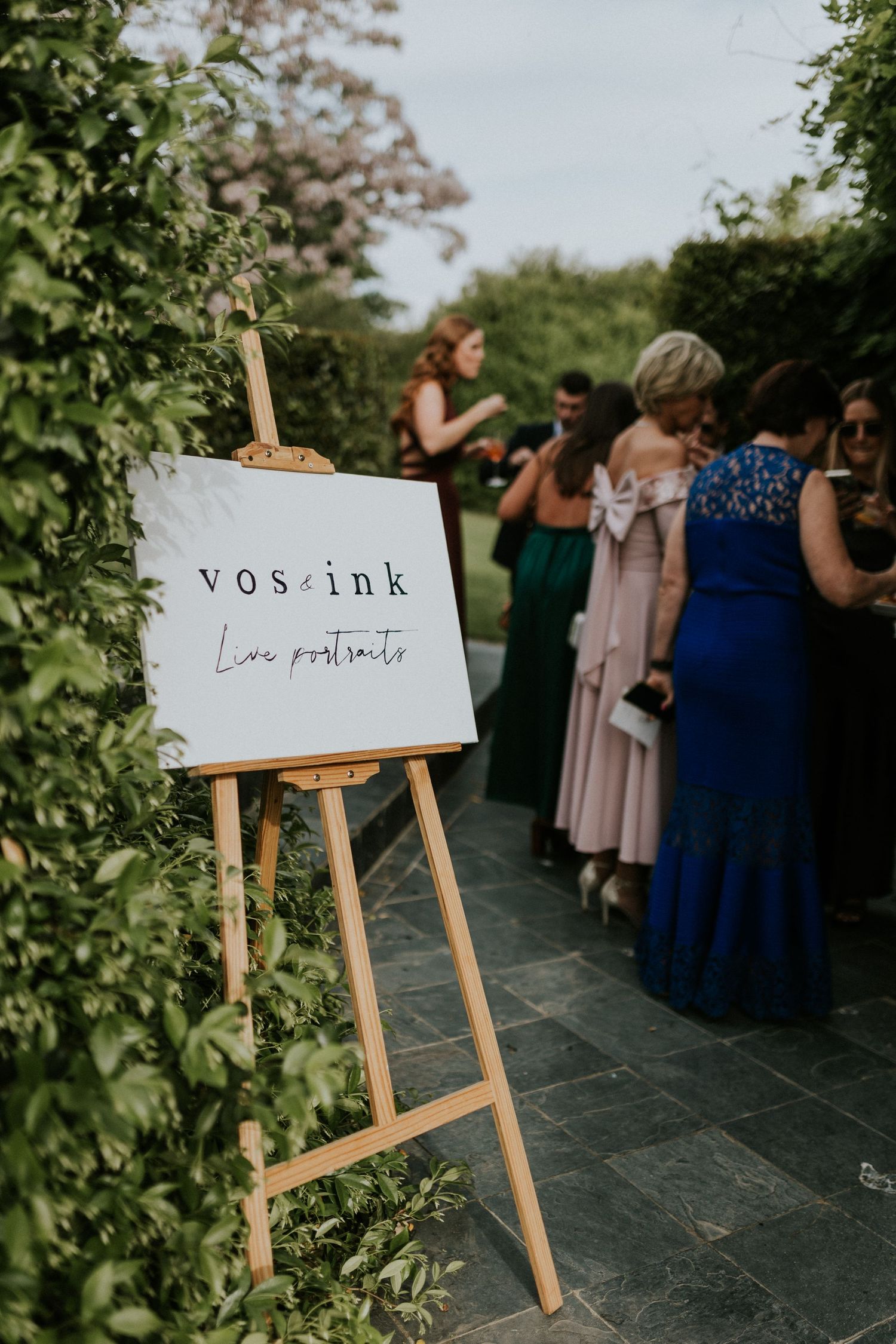 Vos and Ink live portraits sign on wooden easel surrounded by wedding guests in colorful formal attire.