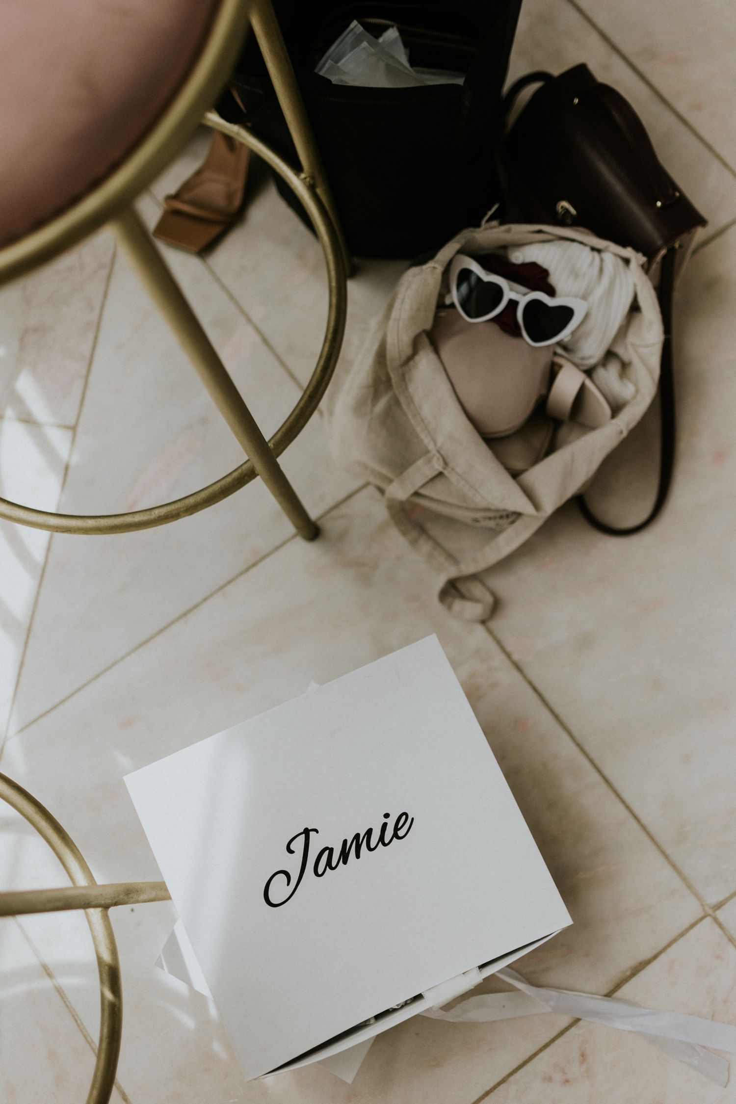 White gift box labeled Jamie on marble floor beside gold chair, sunglasses, and designer handbag.