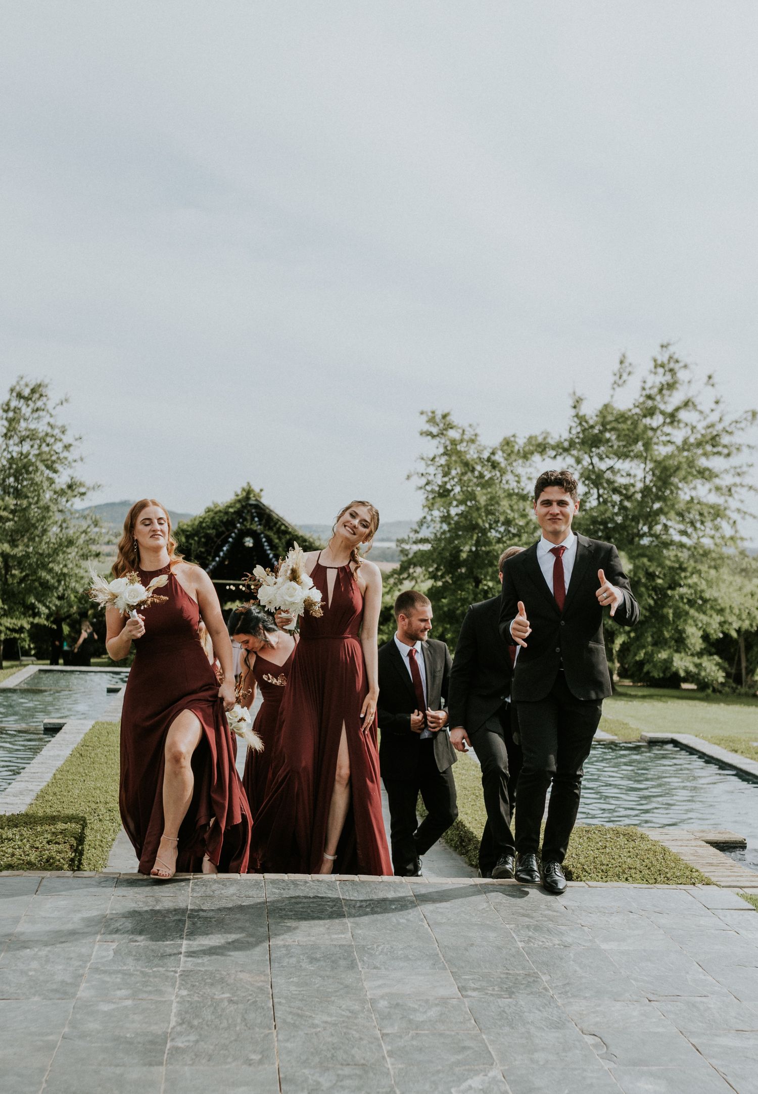 Bridesmaids in burgundy dresses and groomsmen walk together along a garden pathway after a wedding ceremony.