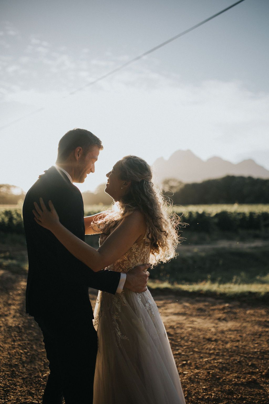 Bride and groom sharing a romantic embrace at golden hour with mountain vineyard backdrop.