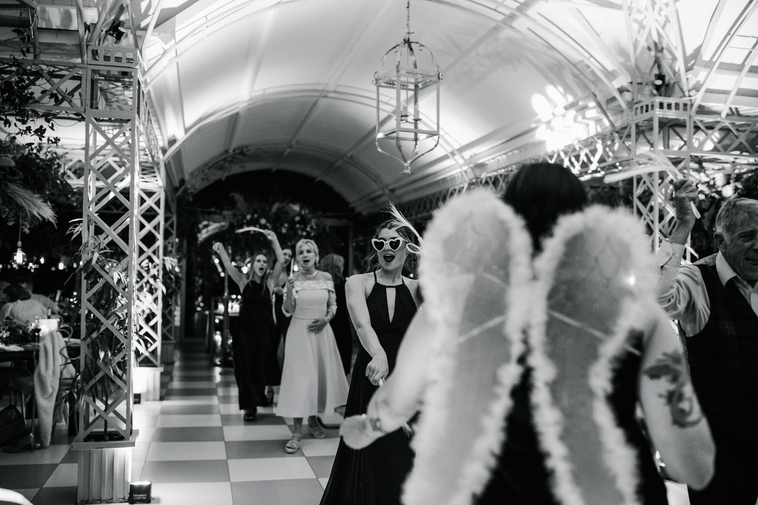 Black and white photo of wedding guests in masquerade masks walking through an elegant arched venue with checkered floors.