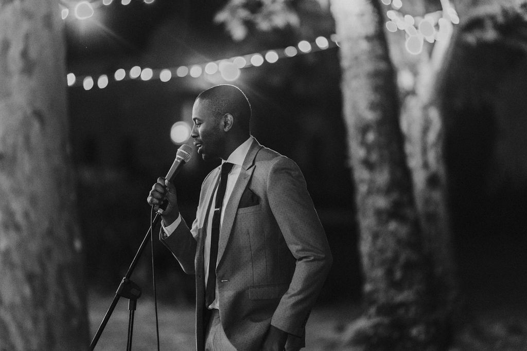 Stylish man in suit speaking at microphone stand during outdoor nighttime wedding celebration.