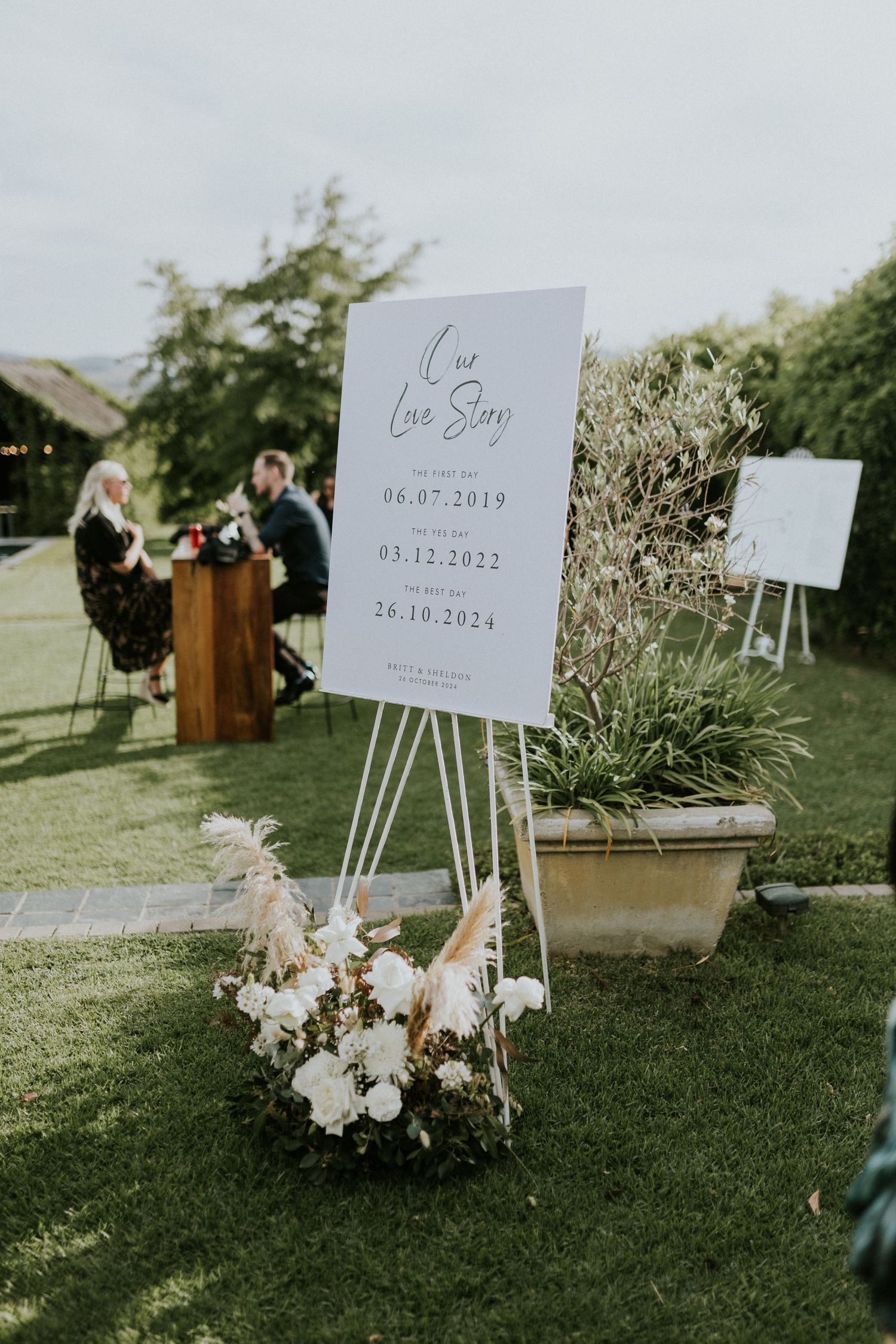 Our Love Story wedding timeline sign on white easel with floral arrangement at outdoor garden reception.
