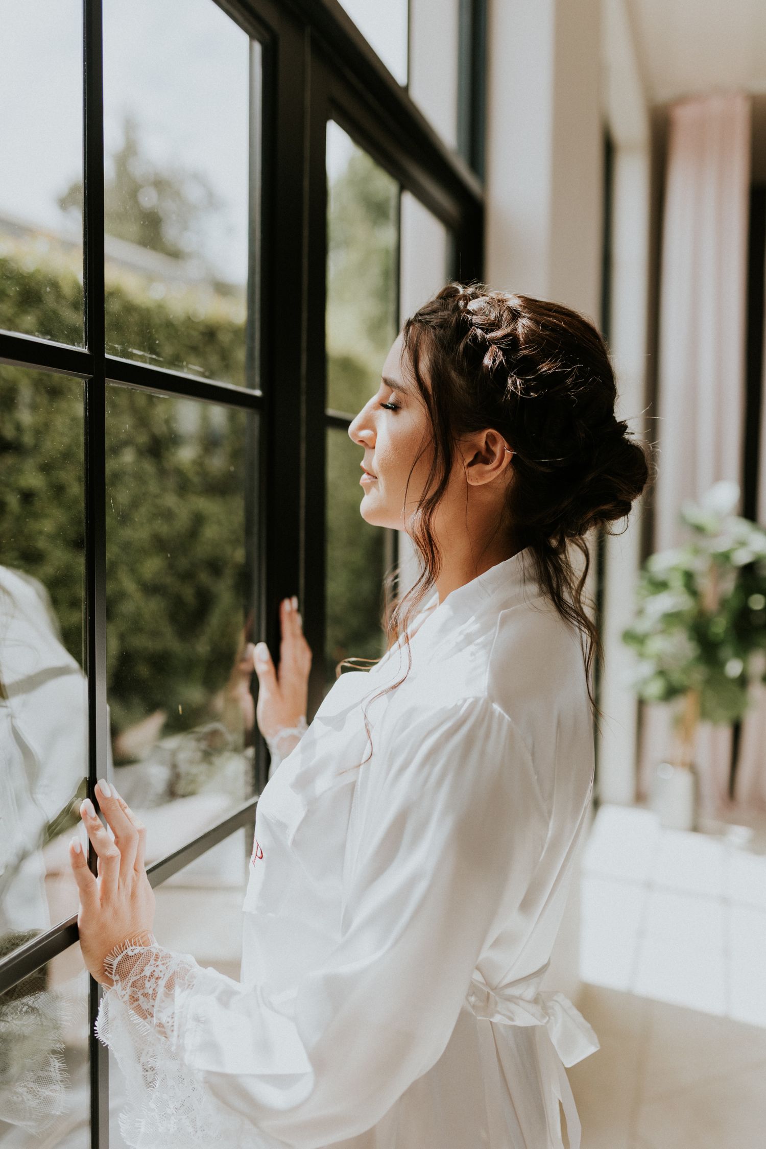Bride in white lace robe with braided updo gazes out large black-framed window.