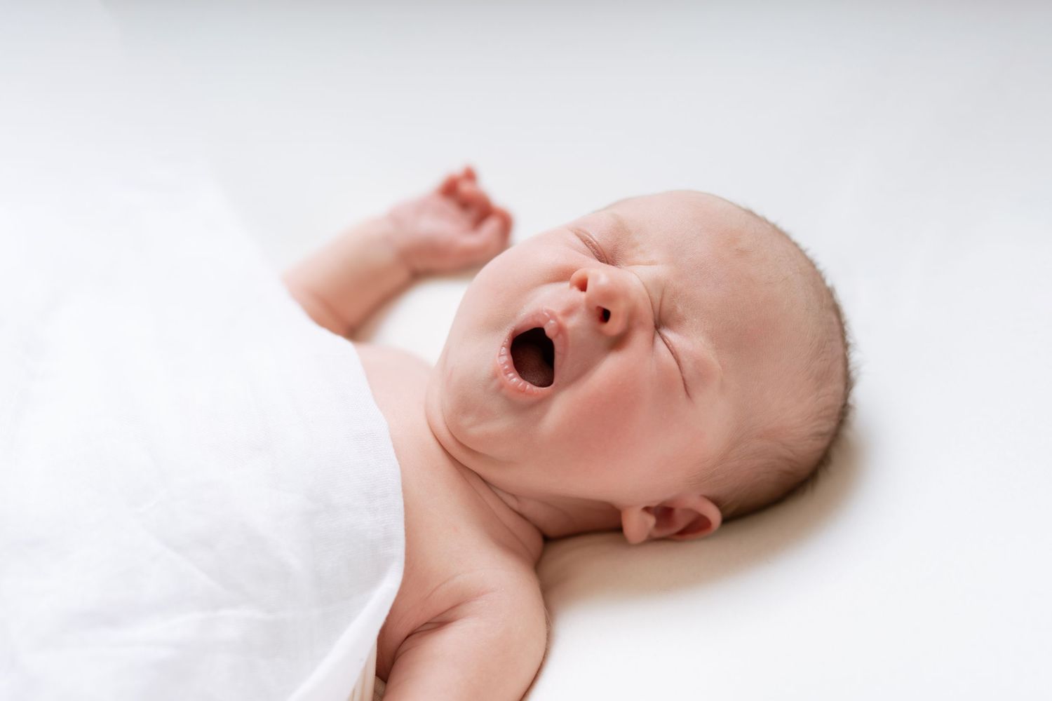 A yawning baby wrapped in white cloth lies against a light background.