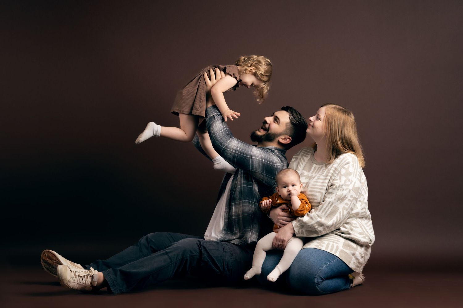 A family plays together on a dark studio backdrop, with one child lifted playfully in the air.