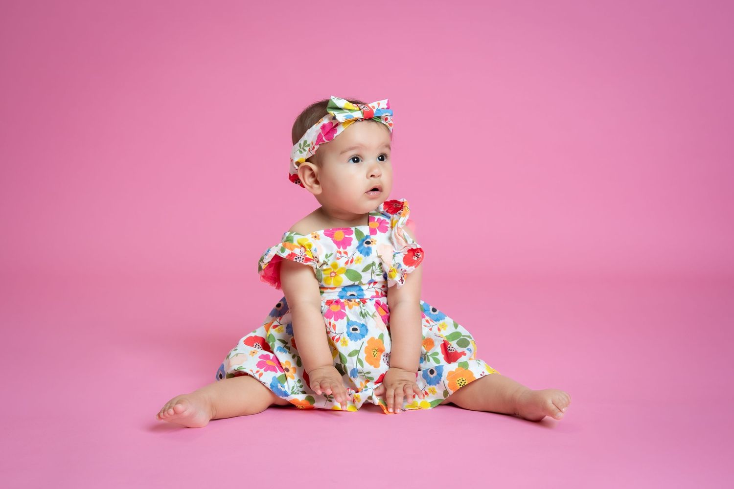 Baby in floral print dress and headband sitting on pink background during studio photography session.