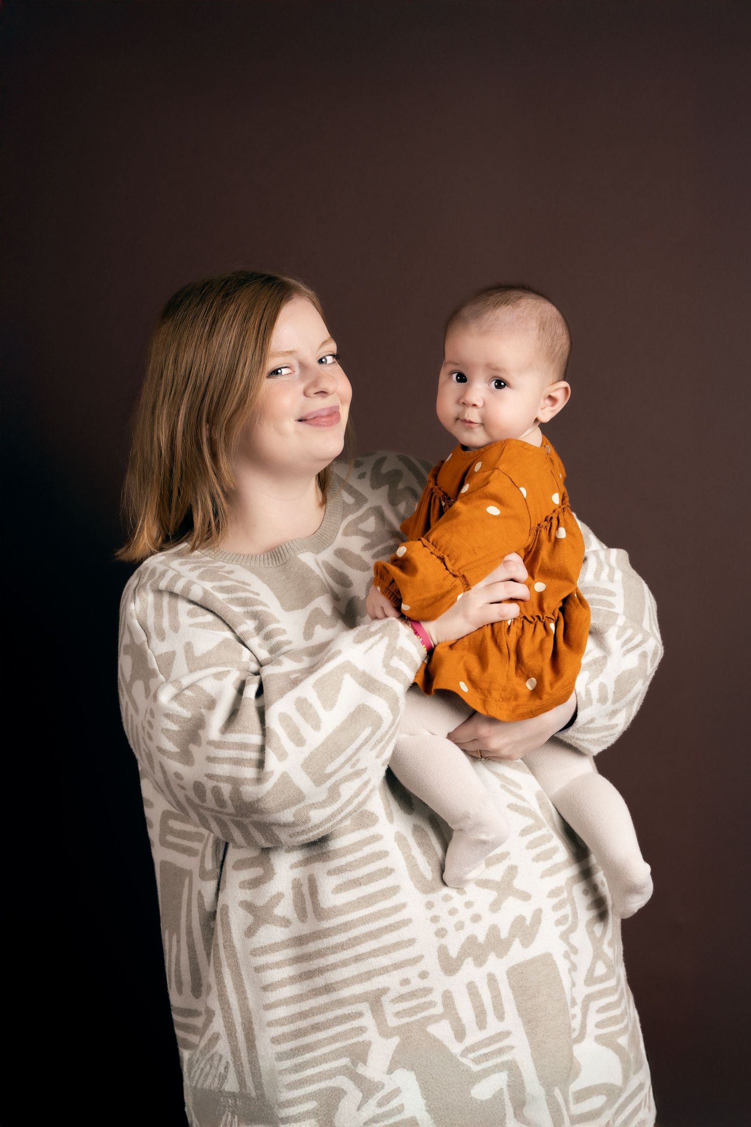 Parent in gray striped sweater shares tender moments with baby wearing orange outfit against dark background.