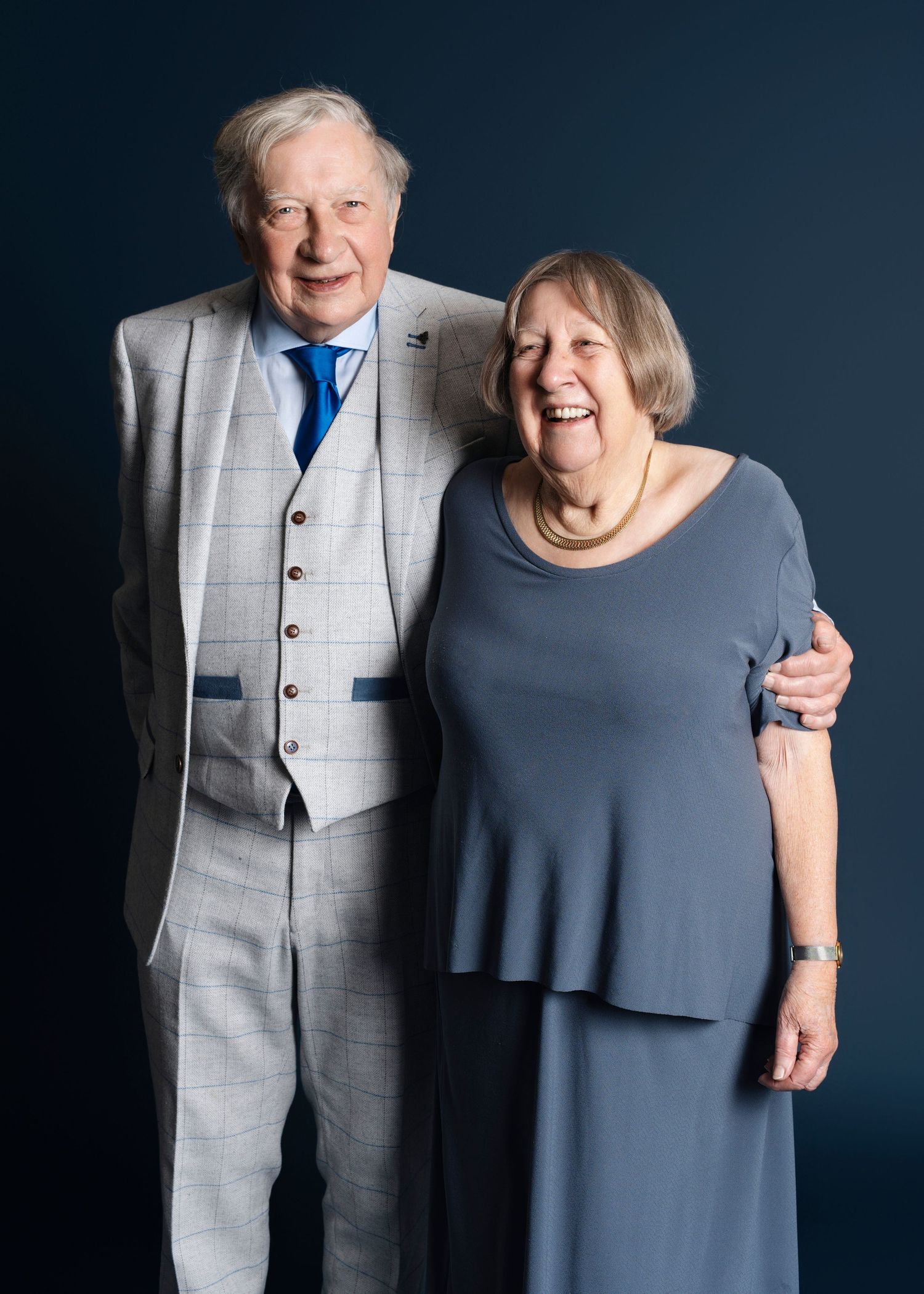 A couple in contrasting outfits of gray suit and navy dress pose together against a dark background in a studio setting.