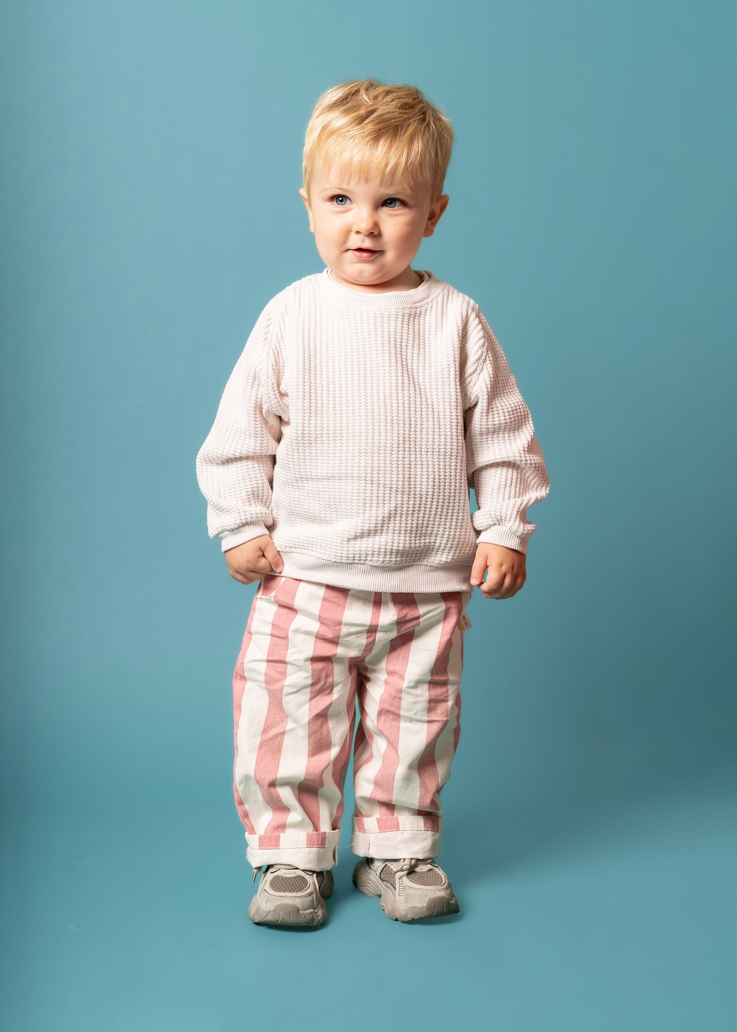 Small child wearing a cream ribbed sweater and pink striped pants stands against a turquoise background in Shropshire photography studio