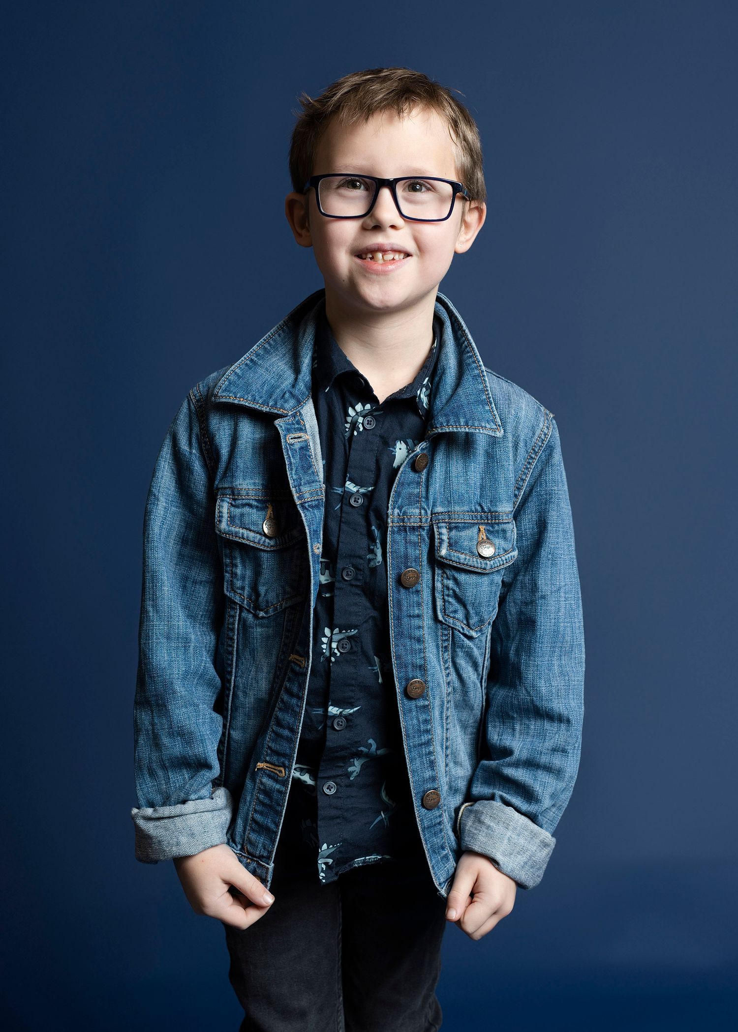 Stylish young person wearing glasses and denim jacket poses against dark blue background with bright smile.
