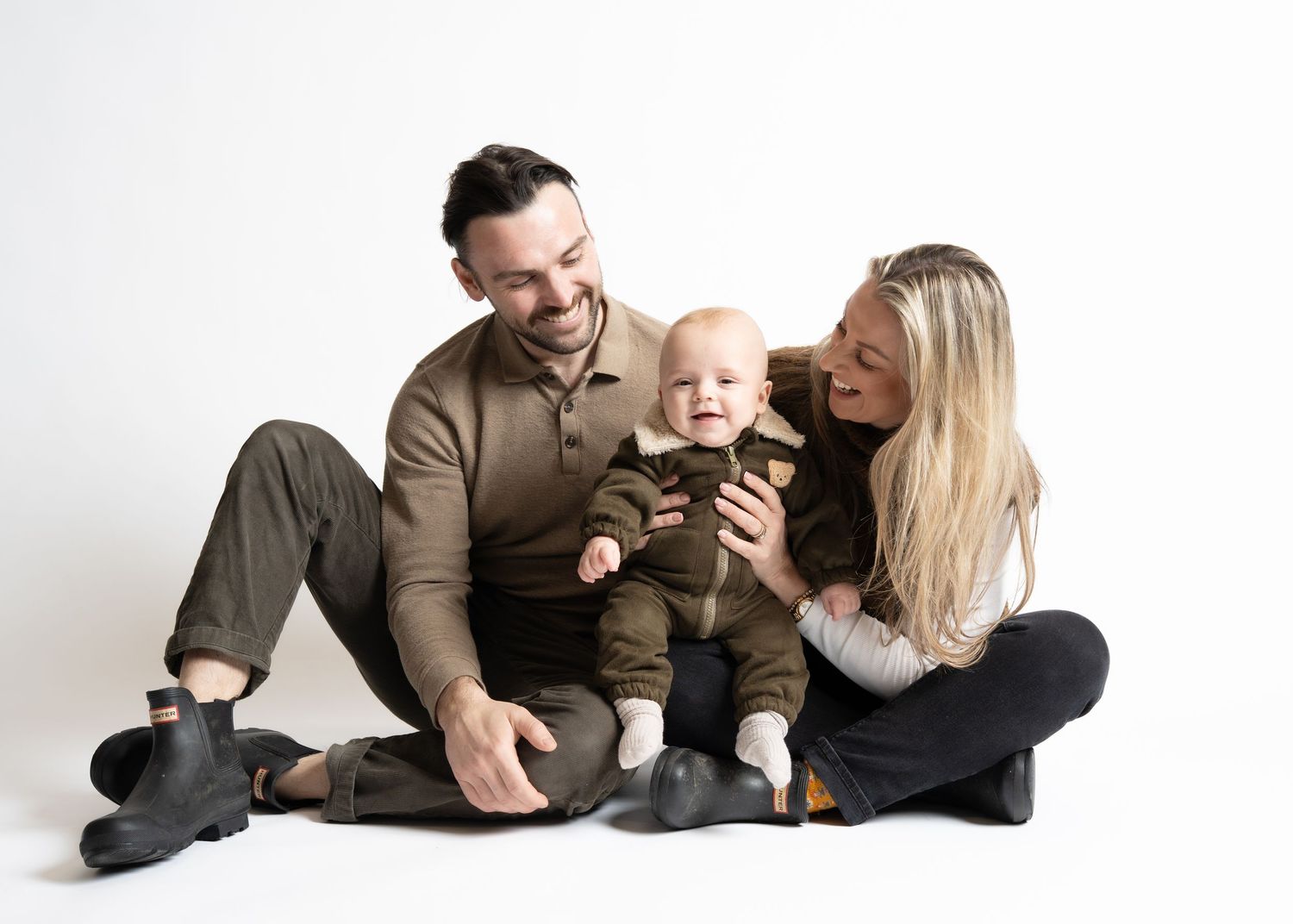 A family sits together on a white backdrop, wearing coordinating brown and black outfits while smiling and embracing.