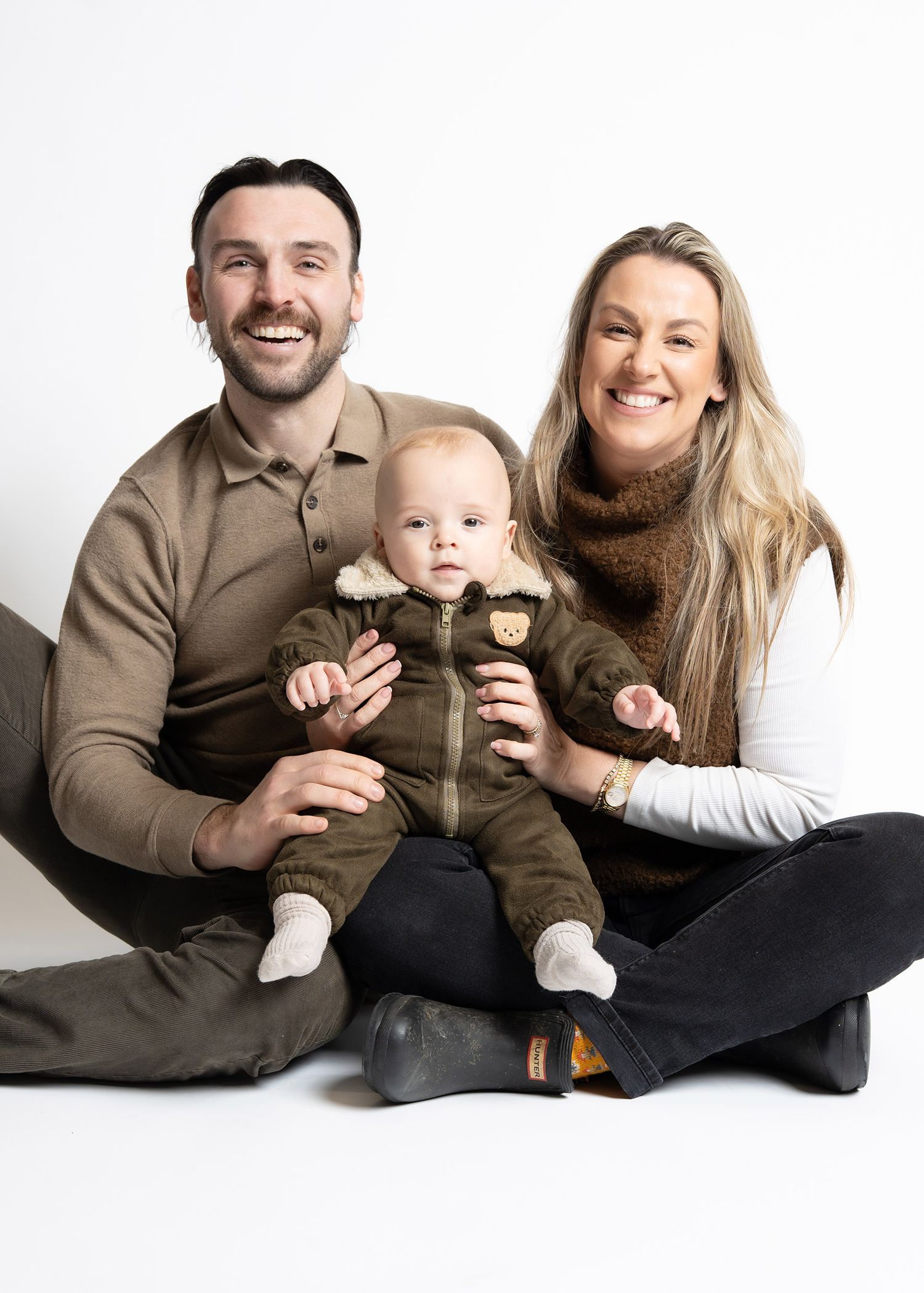 A family sits together on a white background wearing coordinated brown and neutral toned outfits for a portrait session.