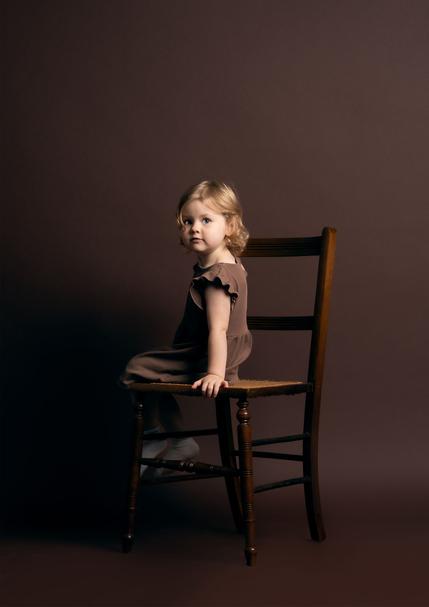 A series of four moody photographs showing a child in a brown dress sitting on a dark wooden chair against a shadowy backdrop.