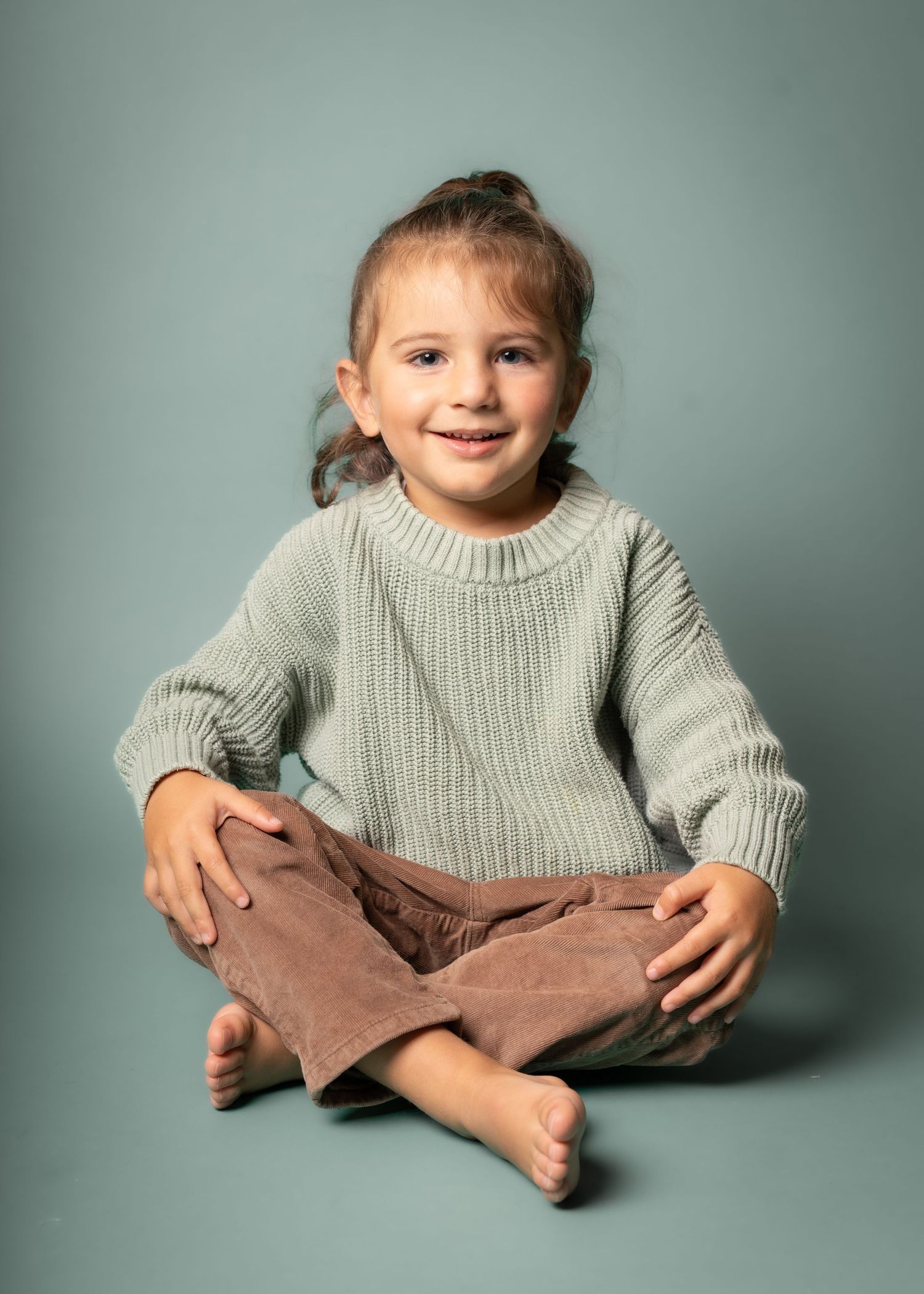 A young child in a cozy gray sweater sits cross-legged on a gray background with a joyful expression.