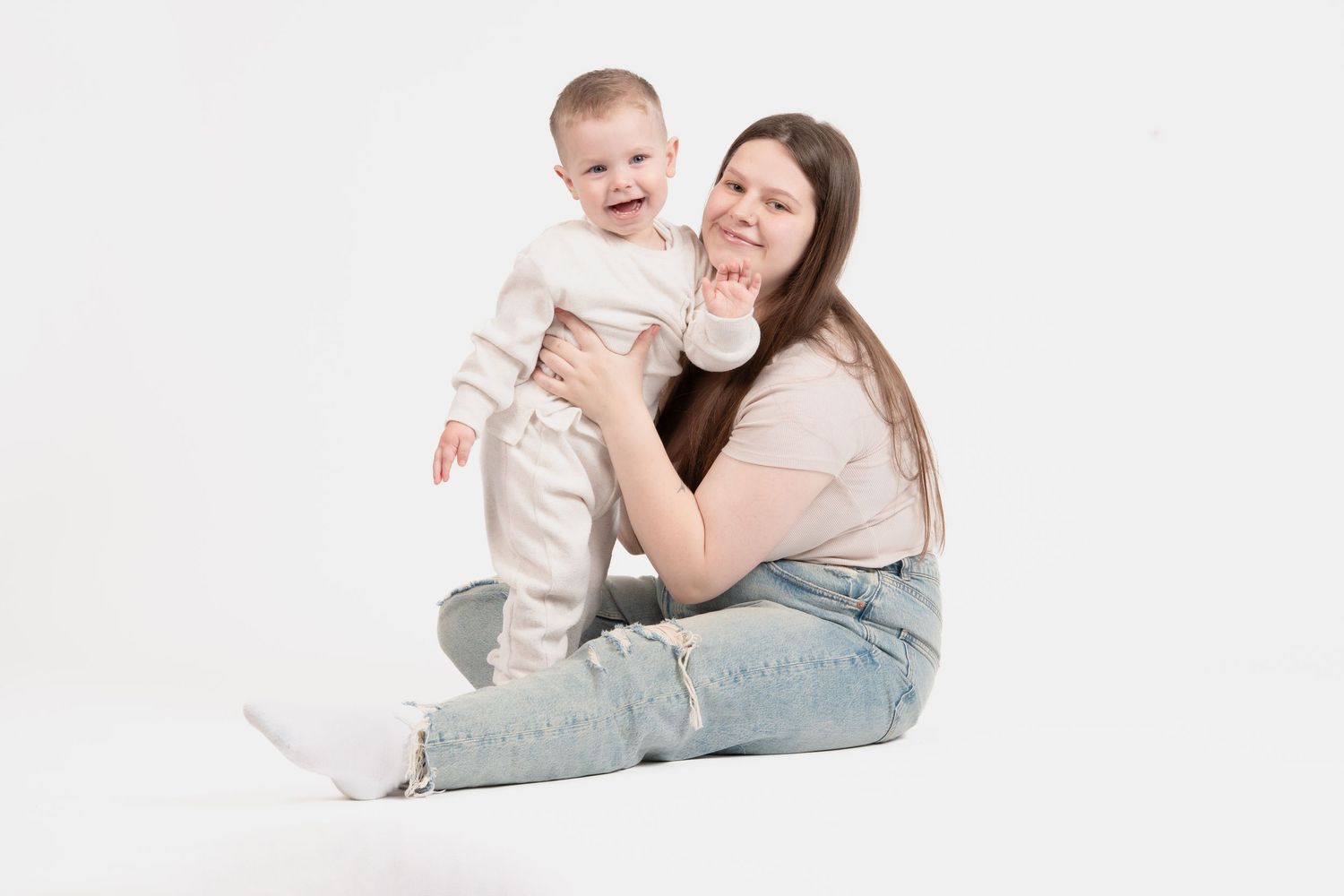 A mother and baby in white outfits play and laugh together on a bright background during a portrait session.