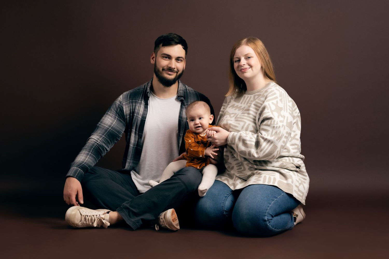 Family sitting together on brown studio backdrop wearing casual winter sweaters and plaid.