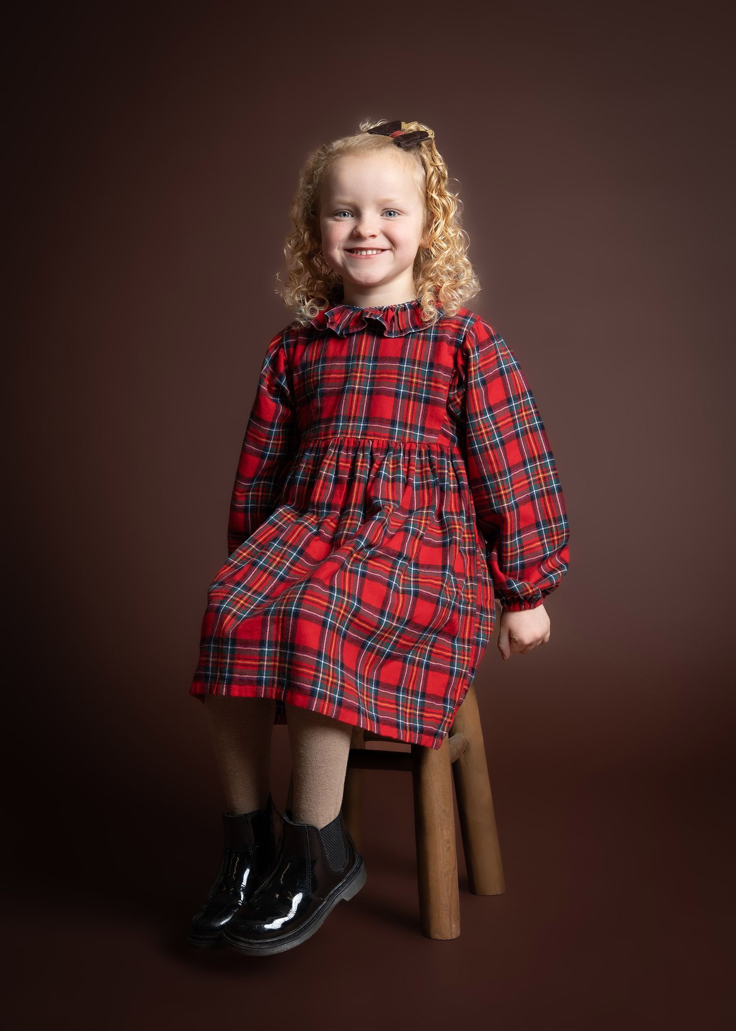 Child in red plaid dress and black shoes sitting on wooden stool against brown background in a studio near Bridgnorth