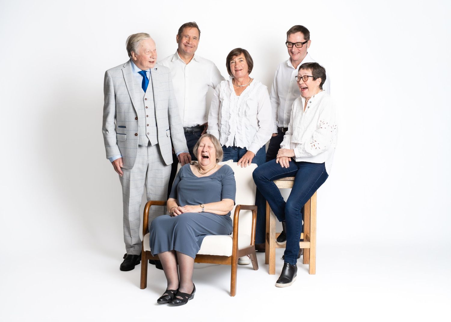 Family portrait series showing group sitting on wooden chairs against white background in casual attire.