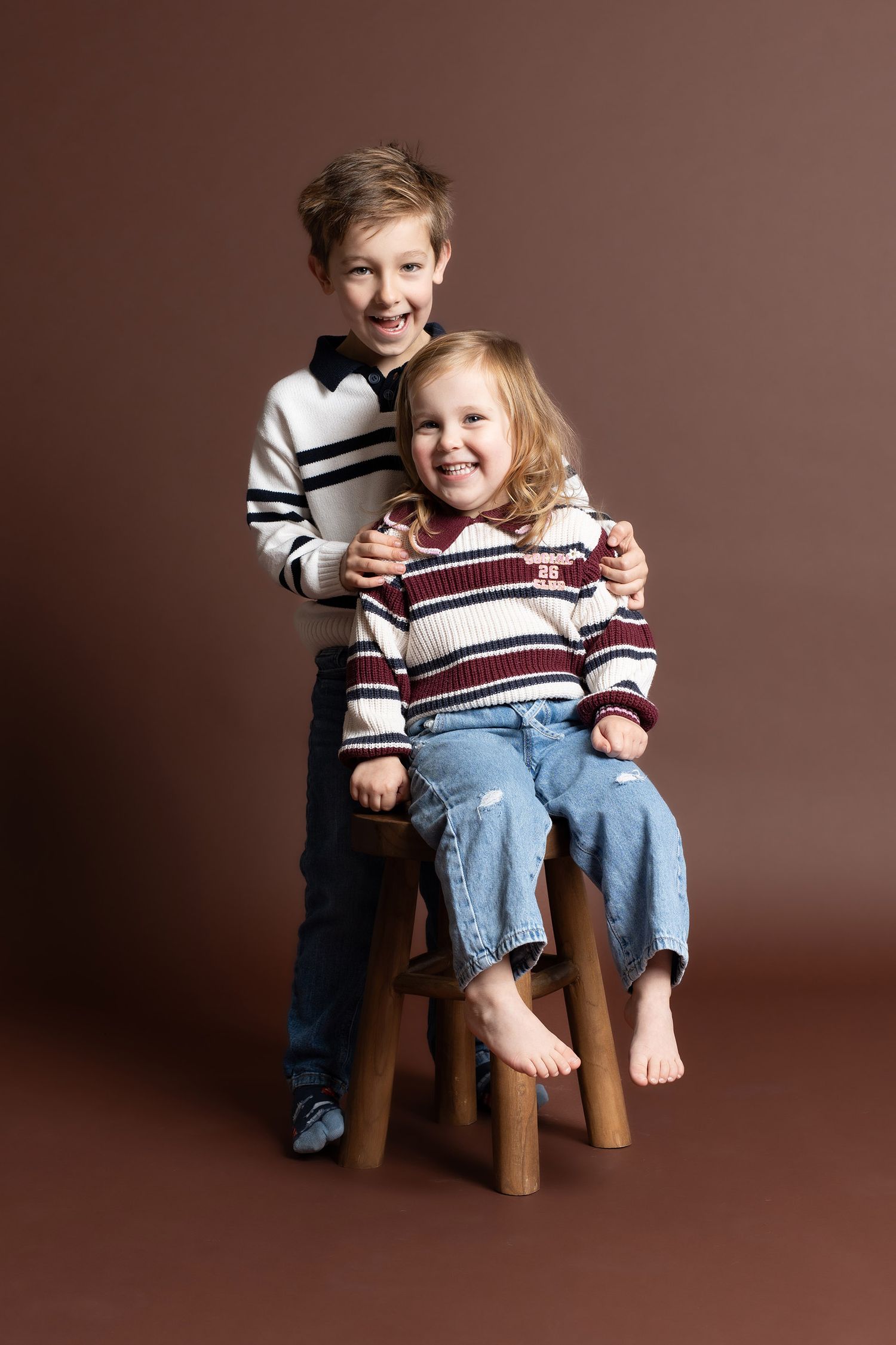 Siblings pose on wooden stool wearing striped sweater and jeans against brown backdrop in studio setting in Telford