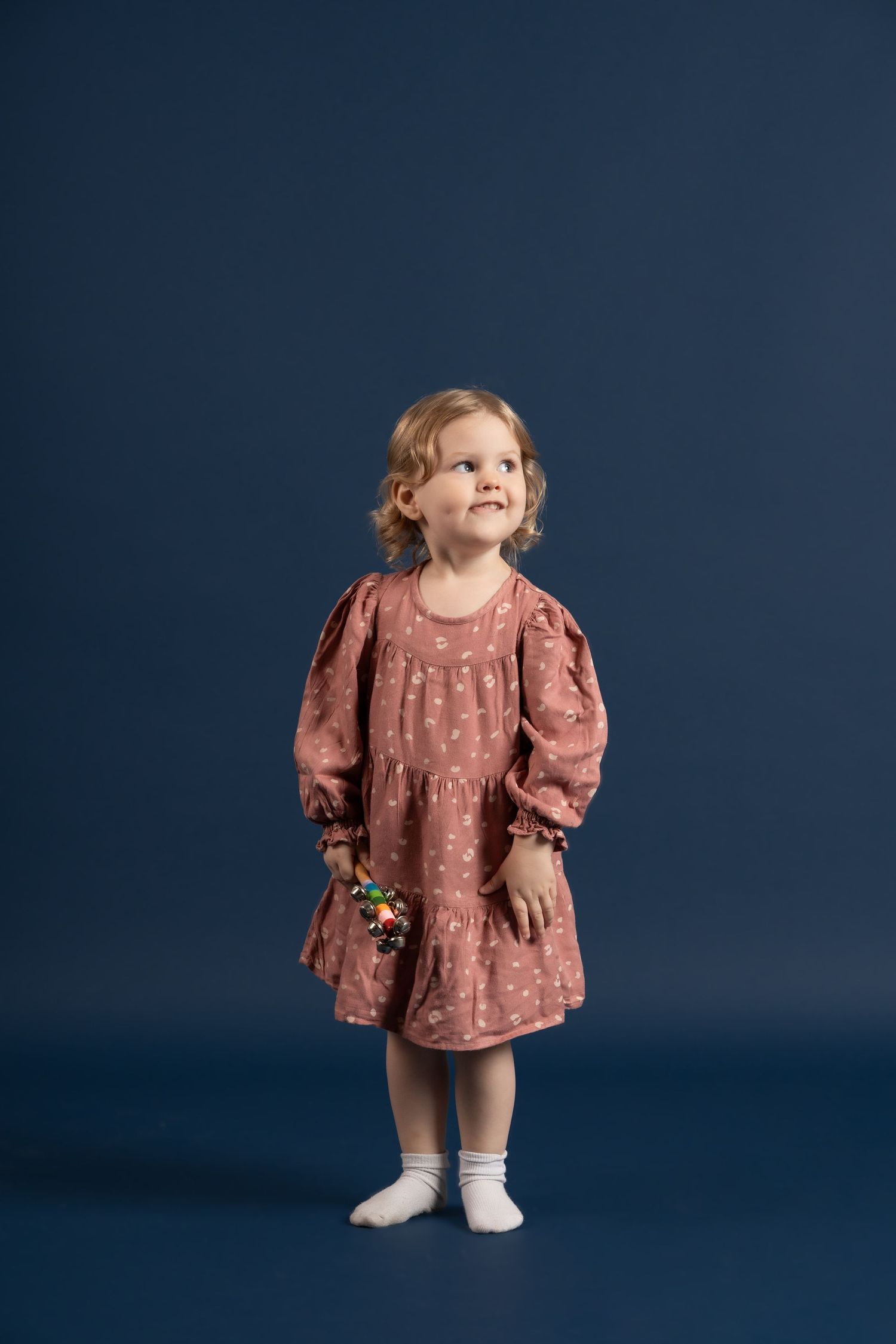 A young child in a dusty rose floral dress and white socks stands against a dark blue backdrop.