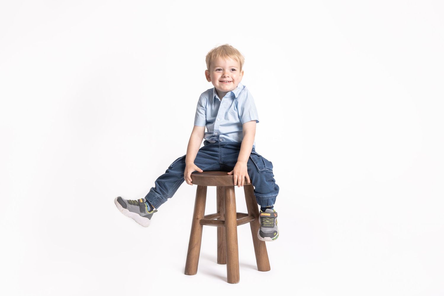 A small child wearing a light blue button-down shirt and jeans sits on a wooden stool against a white background.