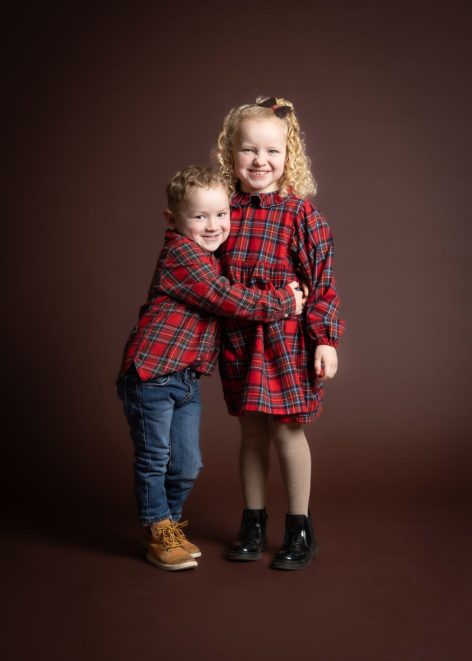 Two children in red plaid shirts and jeans smile while sitting together on a brown background for a sibling photoshoot in Telford
