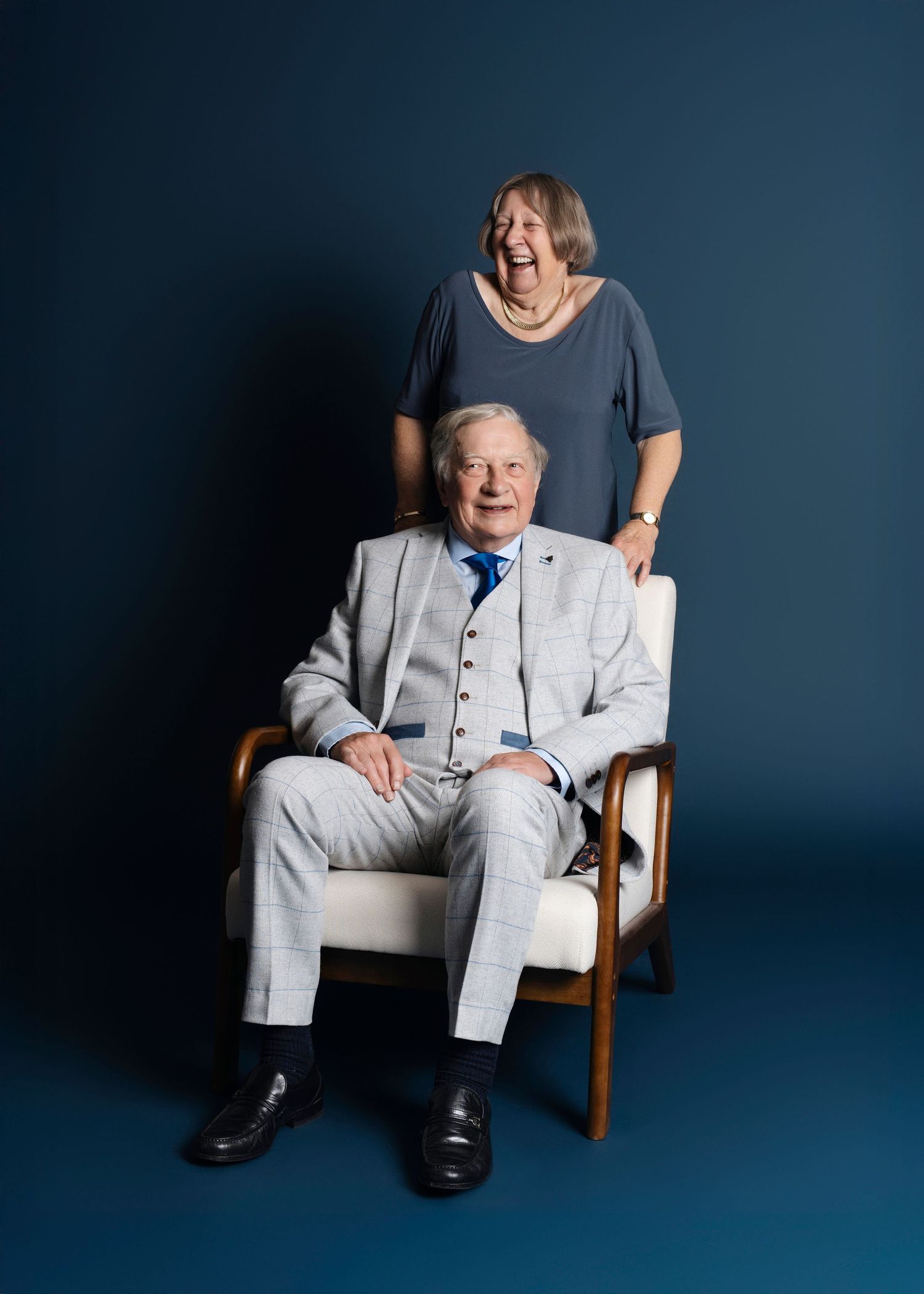 A person in a light gray suit sits in a wooden chair against a dark blue studio backdrop.