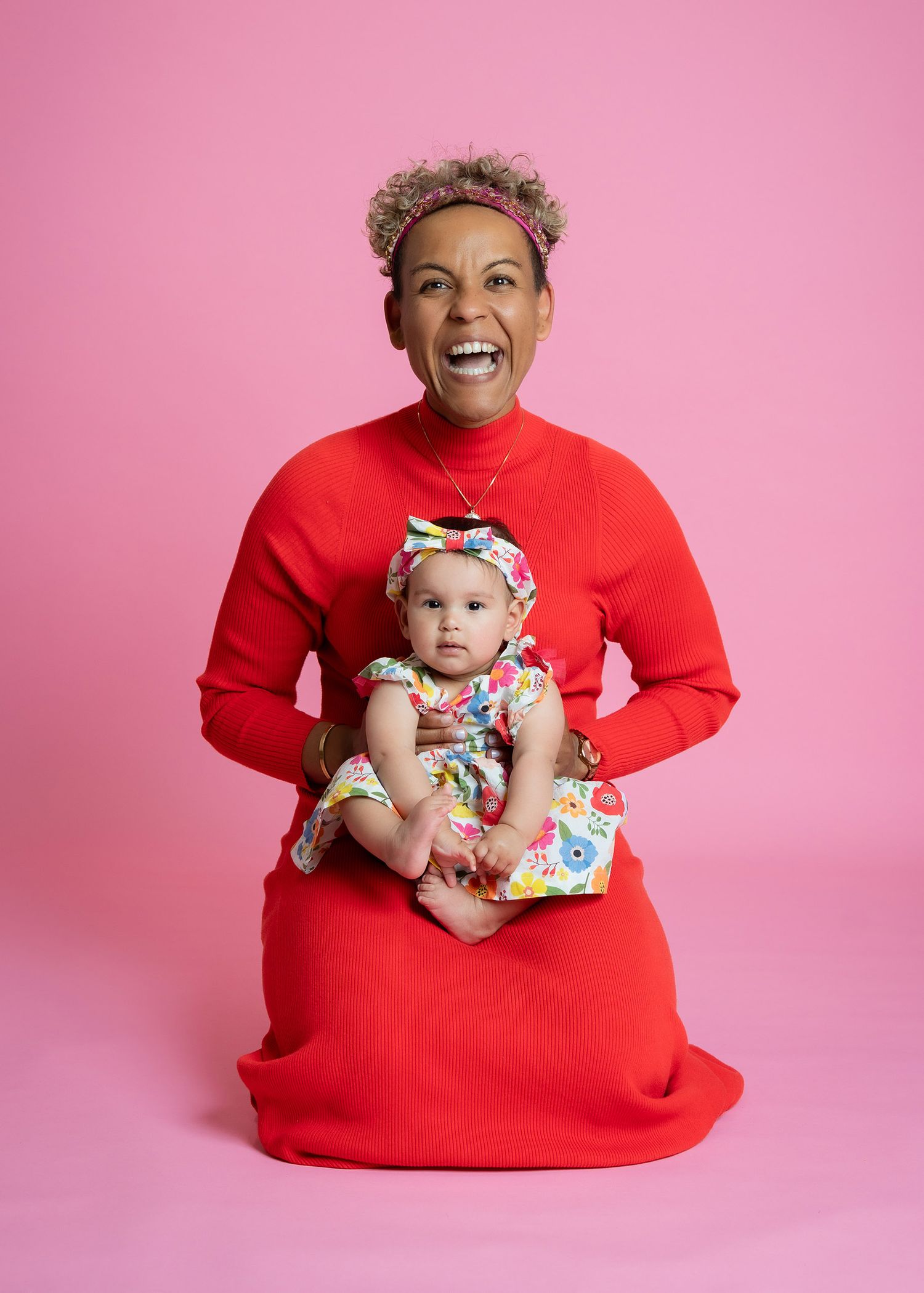 A joyful parent in a bright red sweater dress holds a baby wearing a floral outfit against a pink backdrop.