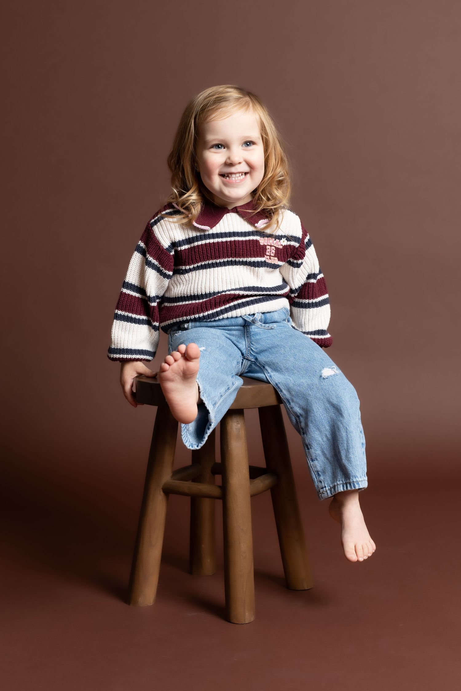 Child in striped sweater and jeans sitting on wooden stool in studio setting with brown backdrop.