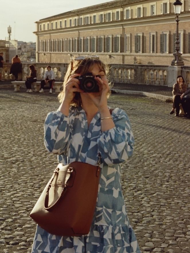 A photographer wearing a blue and white floral dress with a brown leather bag takes pictures on a cobblestone street in Europe.