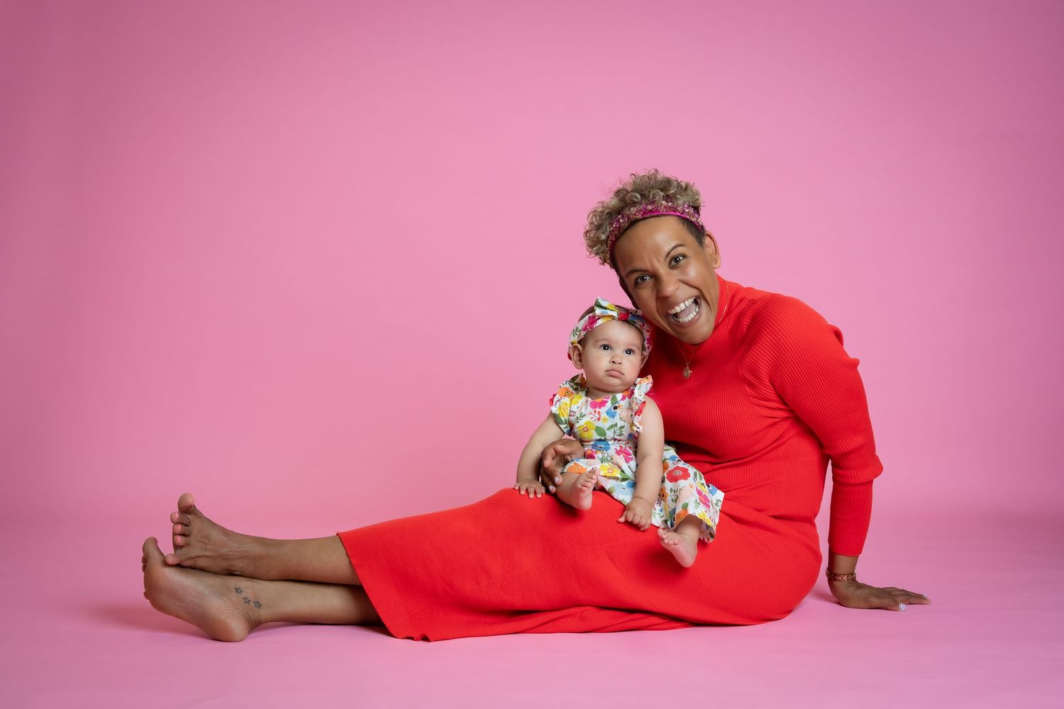 A figure in a red dress reclining with a small child on a pink studio backdrop.