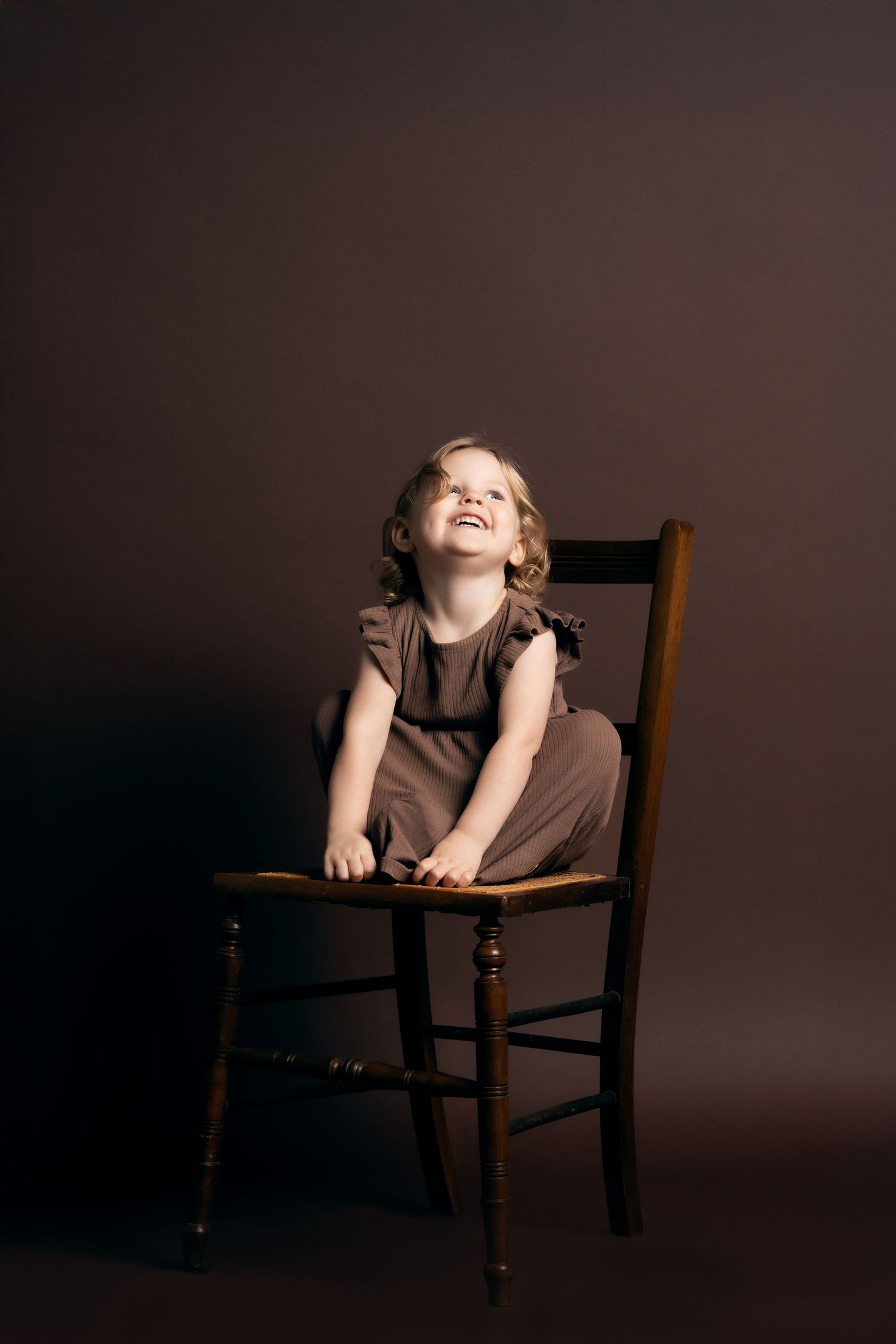 Child with curly hair sits perched playfully on a wooden chair against a dark studio backdrop.