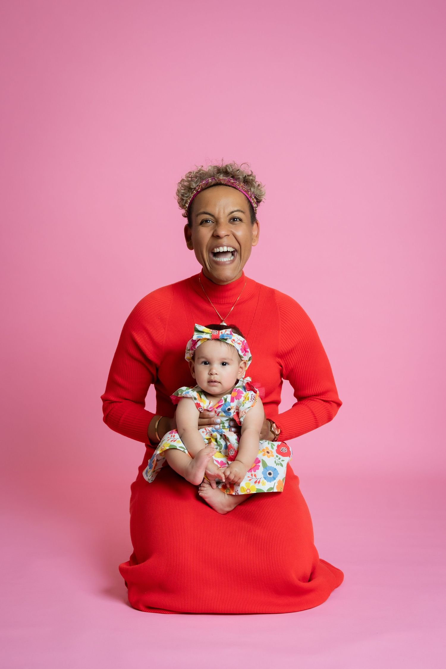 Joyful parent and baby pose in coordinating outfits against a pink backdrop during a studio photography session.