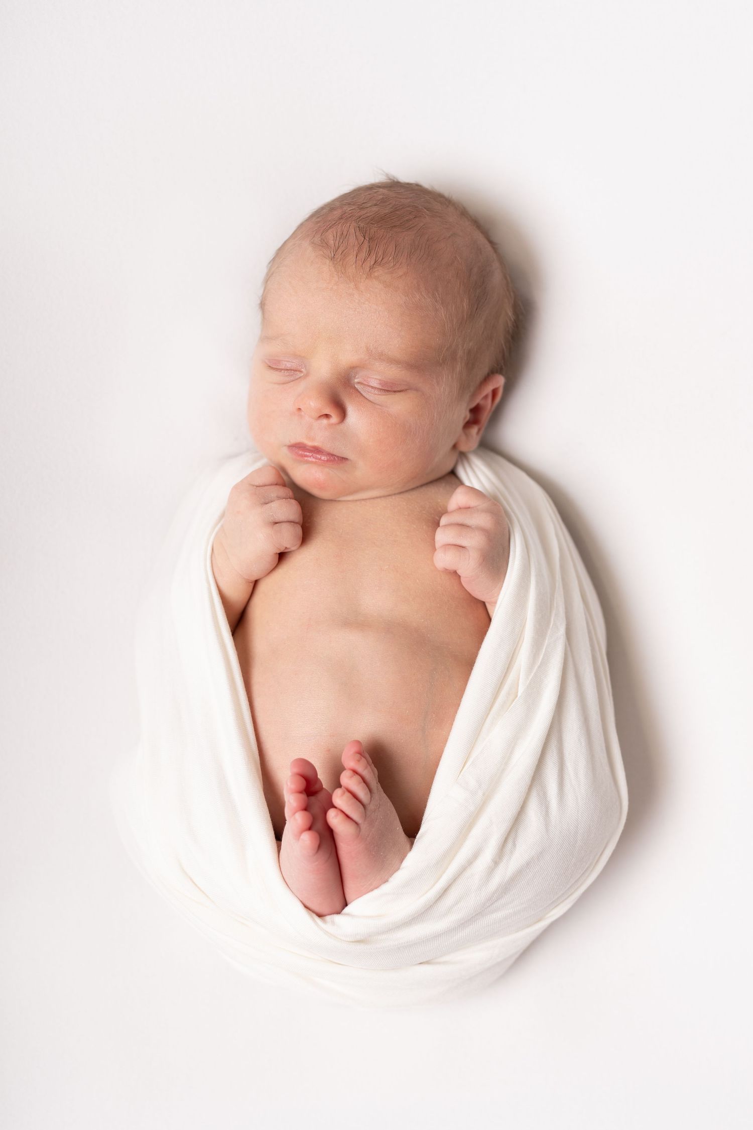 A sleeping newborn wrapped in soft white fabric lies peacefully against a white background.
