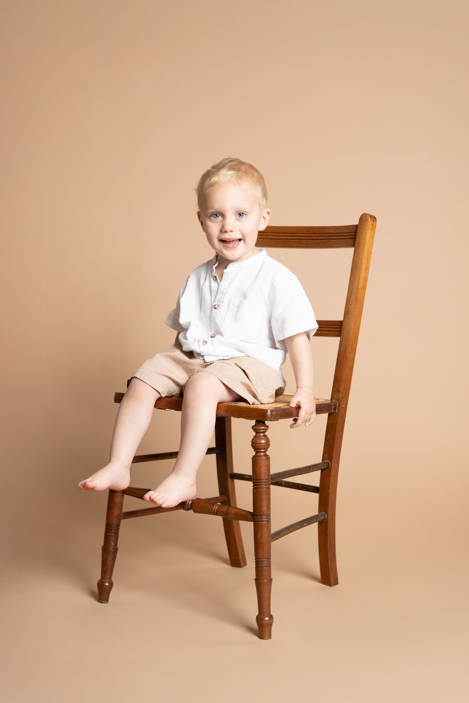 Young child in white shirt and shorts sits barefoot on a wooden ladder-back chair against beige backdrop.