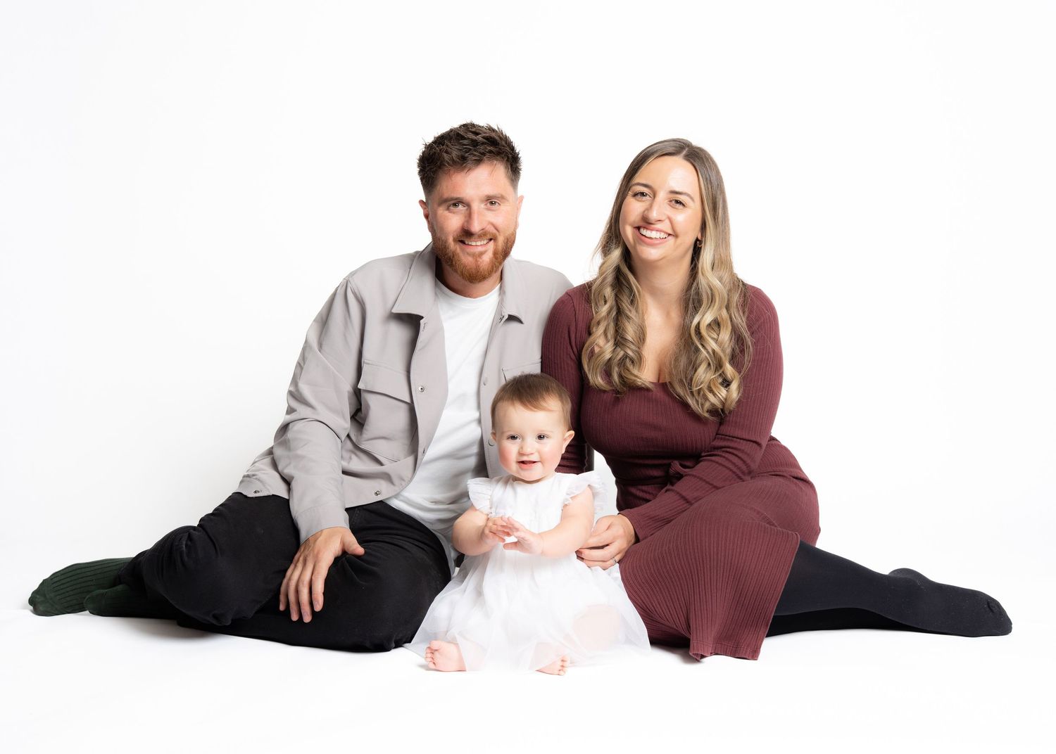 A family in coordinating burgundy and grey outfits poses together on a white backdrop for professional photos.