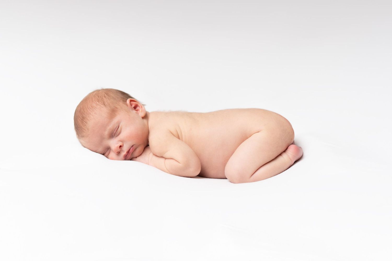 A sleeping newborn baby lies on a soft white background in a curled fetal position.