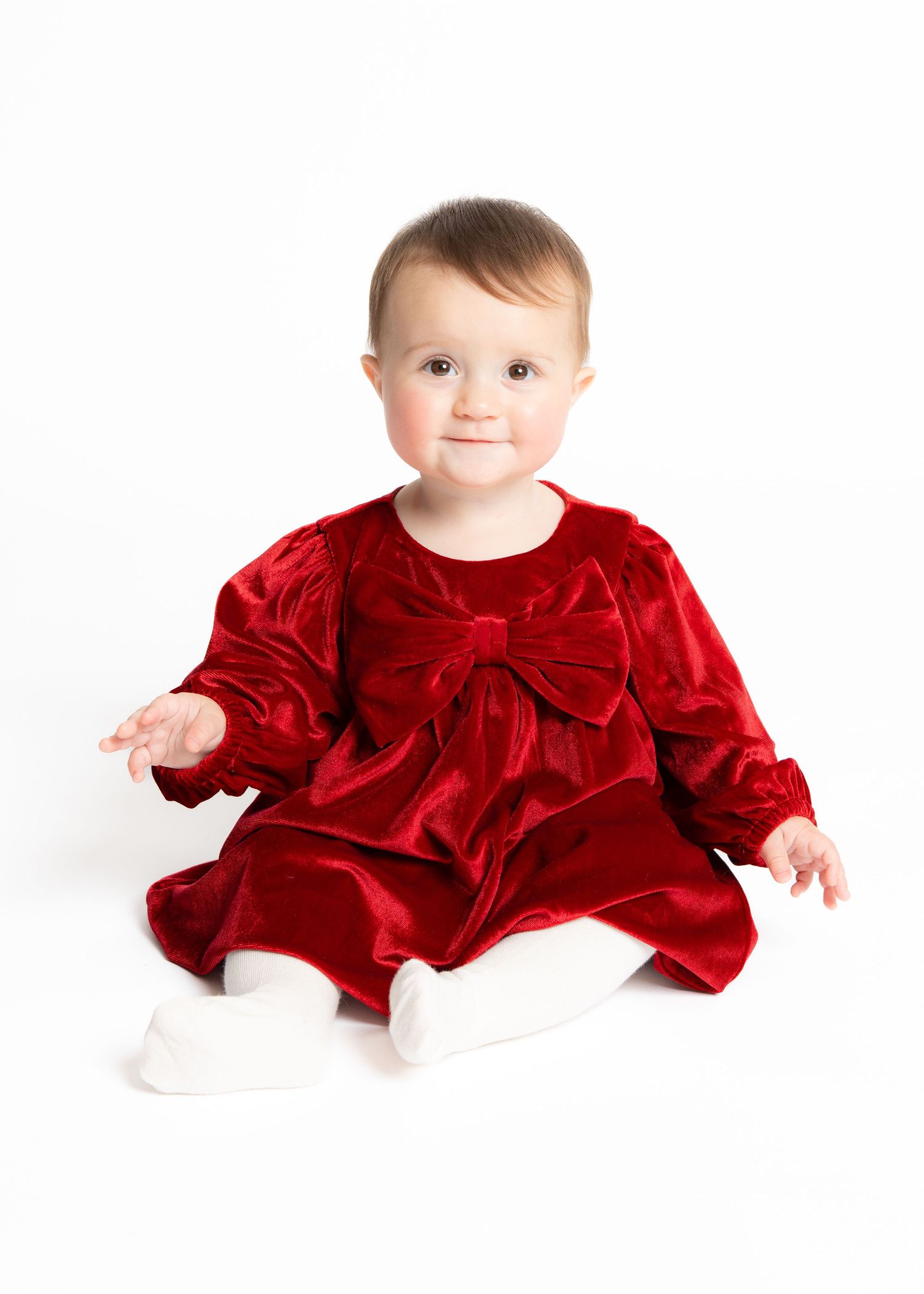 Baby wearing festive red velvet dress with bow detail and white socks sits against white background.
