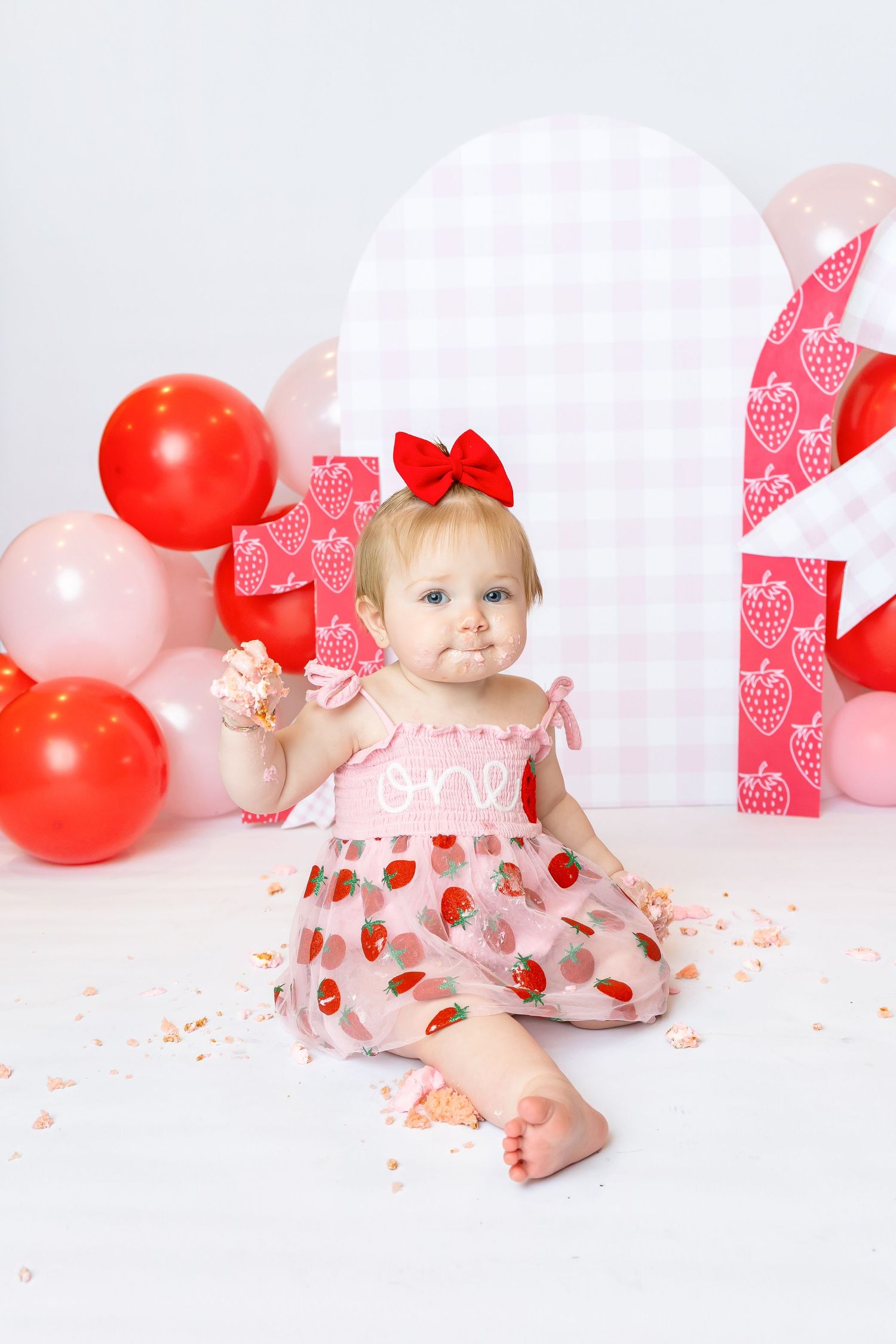 Baby wearing pink strawberry dress sits with hand full of cake grinning during 1st birthday photoshoot with Annie Robinson Photography.